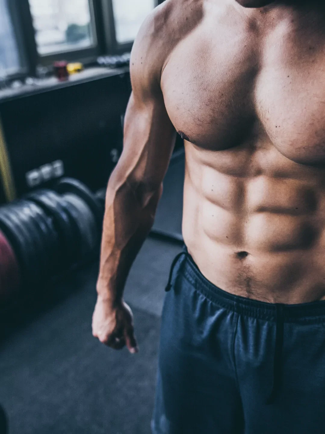 Close-up of a muscular man's upper body showing defined abs, chest, and arm in a gym with weights and windows in the background.