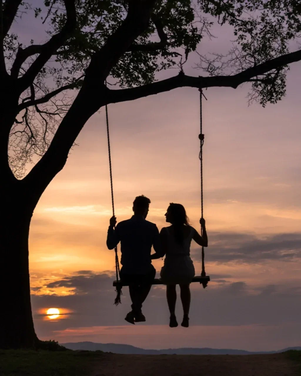 Silhouetted couple sitting on a swing hanging from a tree at sunset.