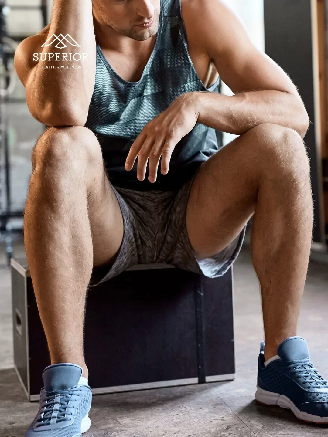 A man in athletic clothing, sitting on a black box in a gym, resting with one hand on his knee and the other on his face, with a