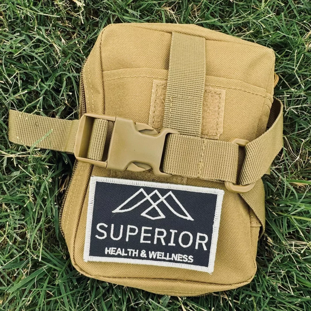 A tan tactical nylon pouch with a buckle strap, placed on grass, featuring a patch that reads 'SUPERIOR HEALTH & WELLNESS' with a mountain logo.