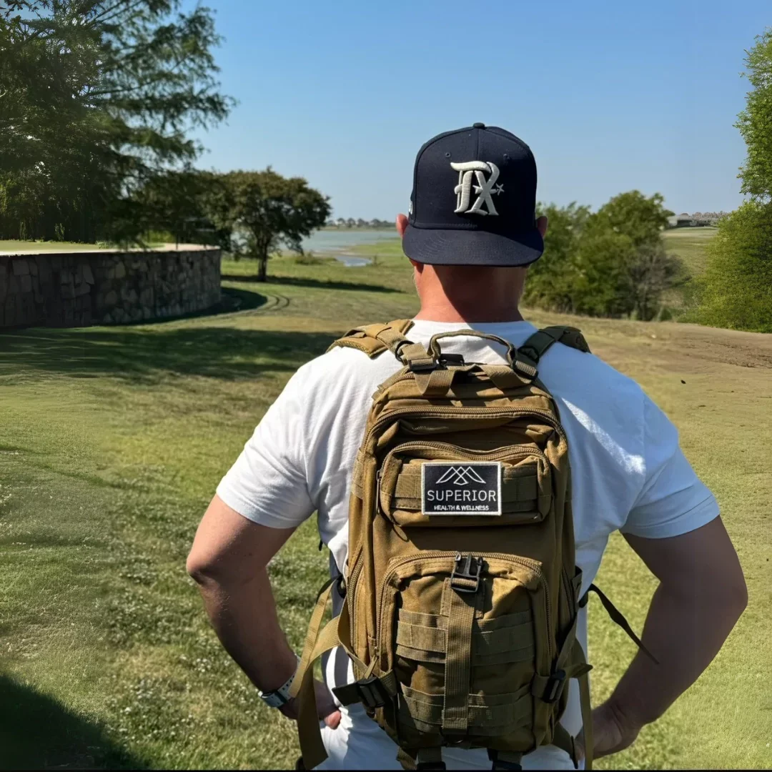 A man wearing a black baseball cap, a white t-shirt, and a tan backpack stands outdoors on a grassy area, looking at a body of water surrounded by trees on a sunny day.
