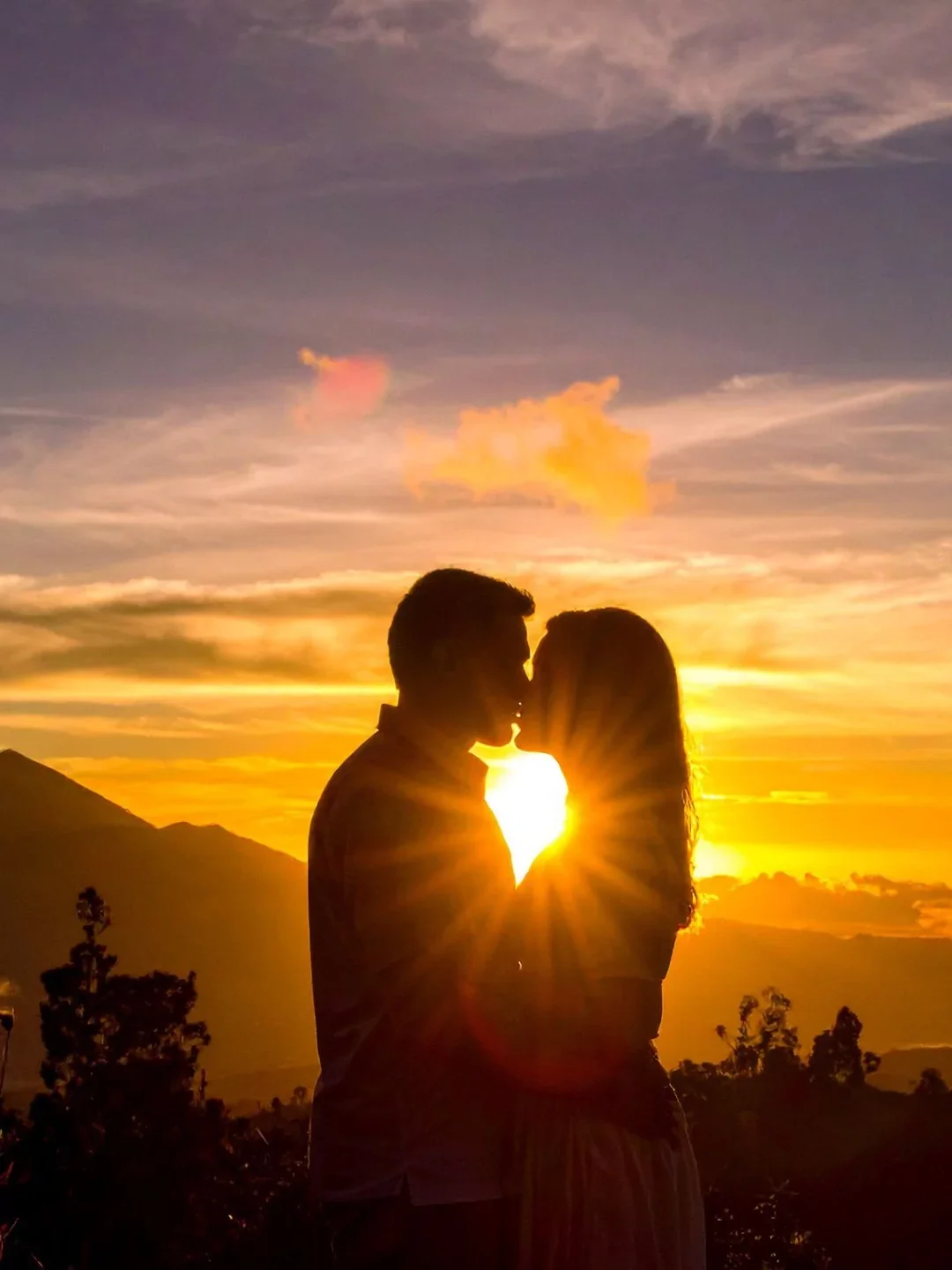 Silhouette of a couple kissing during sunset with mountains and trees in the background.