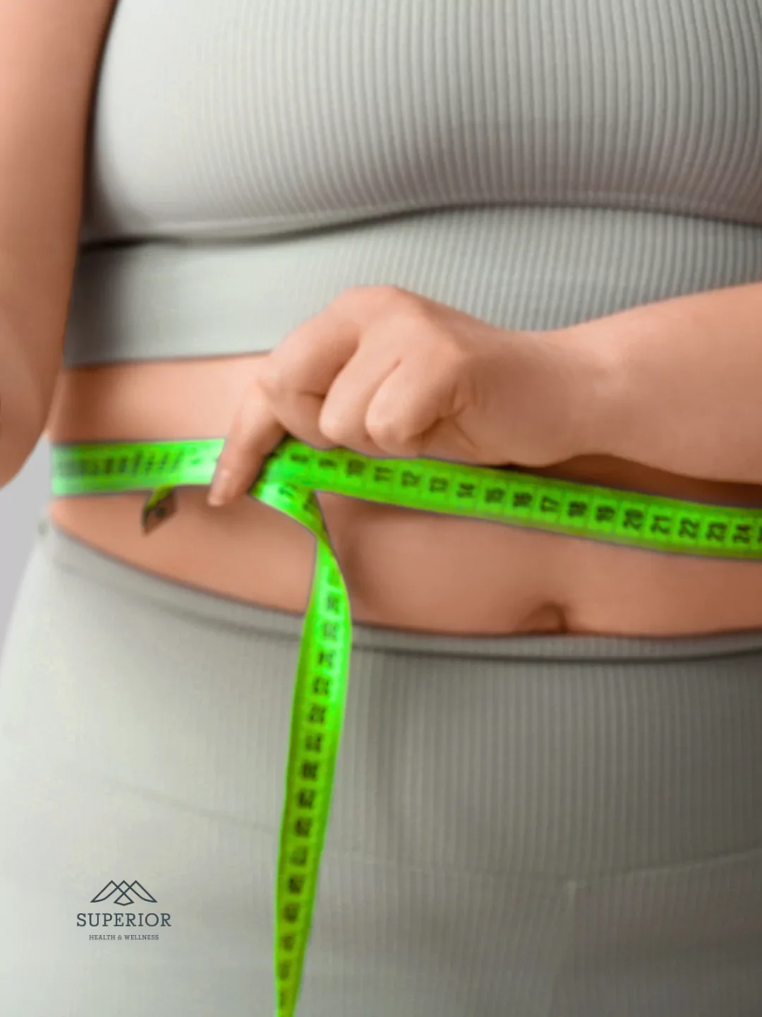 Person measuring waist with a green measuring tape, wearing light gray athletic clothing, promoting health and wellness.