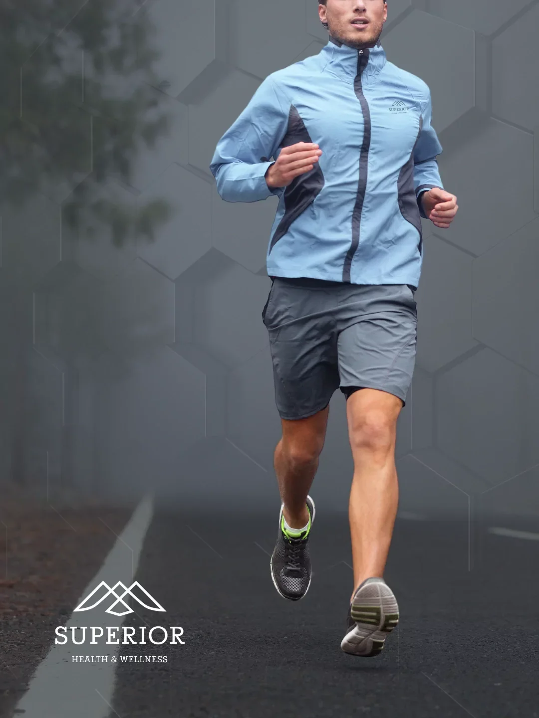 A man running outdoors on a foggy day, wearing a light blue athletic jacket and gray shorts, with a logo that says SUPERIOR on the jacket, on a road with a painted white line.