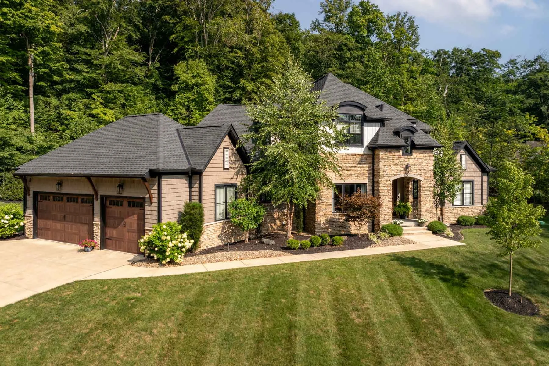 Custom home featuring a stone and wood exterior, black roof, and a well-landscaped front yard with a circular driveway.