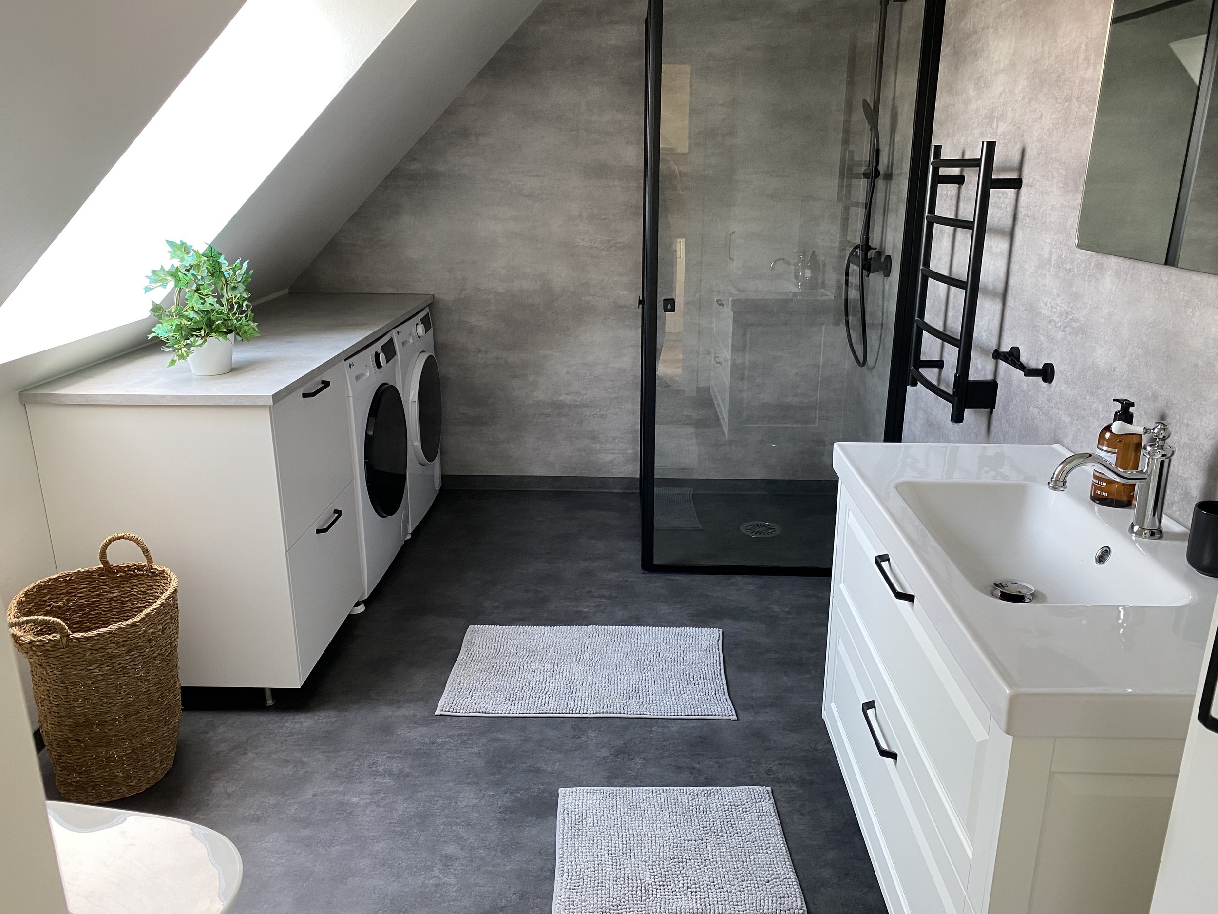 Moderne badrum med tvättmaskin, torktumlare, tvättbänk, badrumsspegel, handfat, dusch med glasvägg, vädringsfönster och inredning i ljusa och mörka toner.