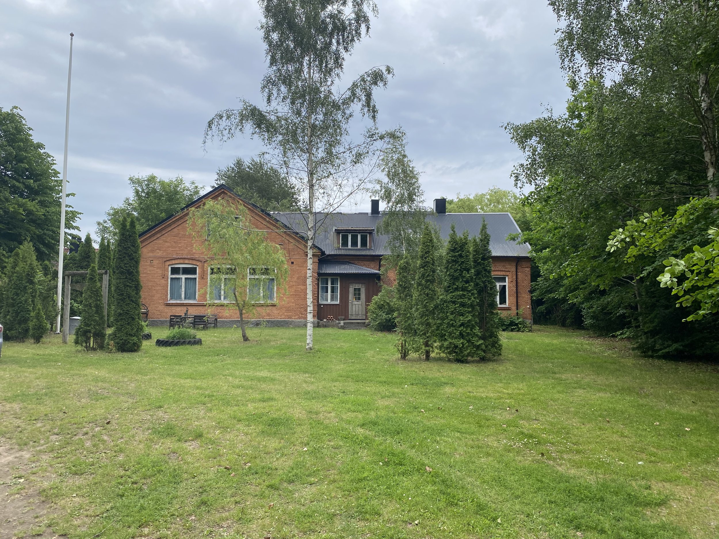 En röd tegelvilla omgiven av gröna träd och en gräsmatta, med en blå himmel med moln.