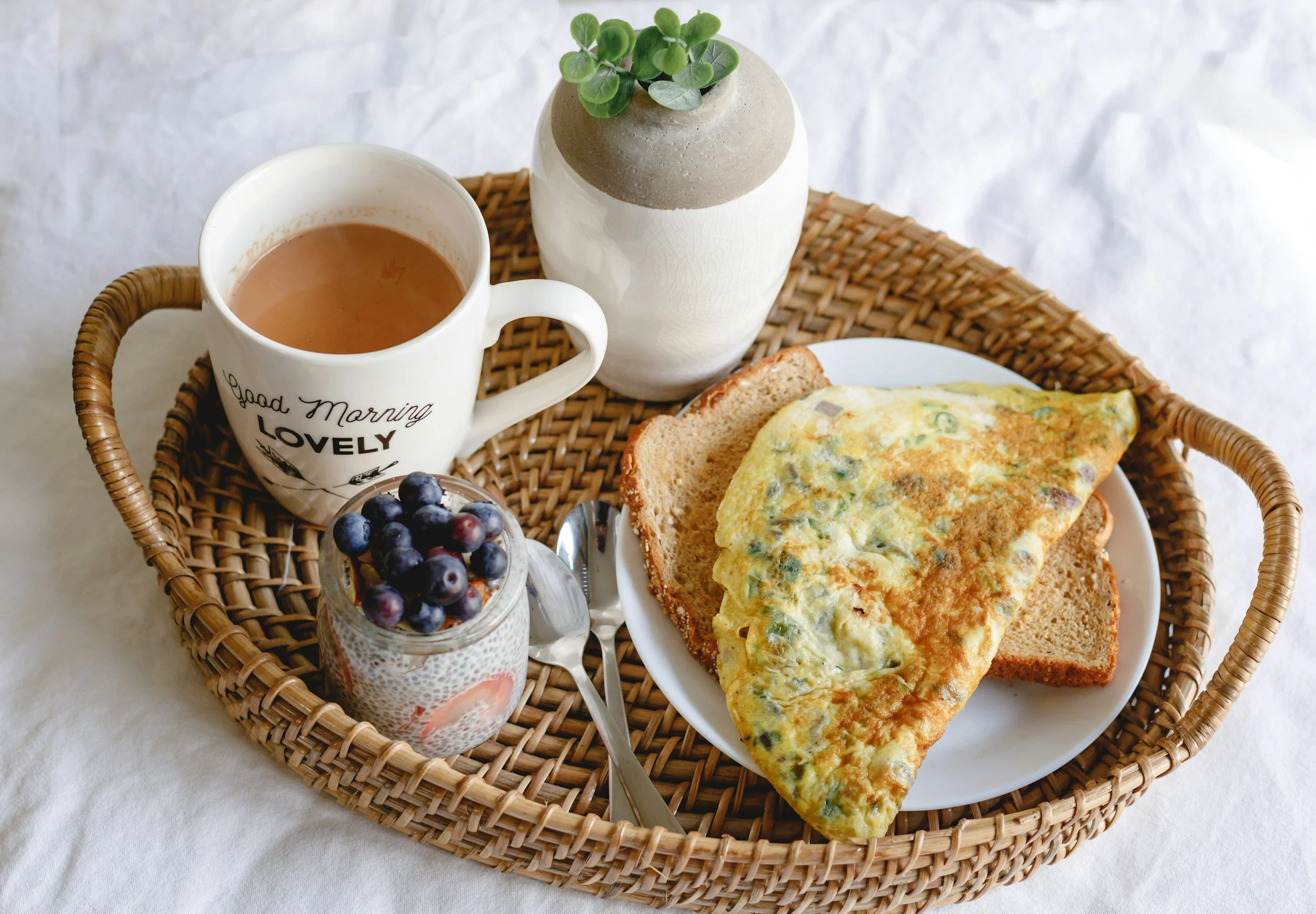 Un petit-déjeuner sur un plateau en osier contenant une tasse de thé ou café, un petit pot de yaourt avec des myrtilles, une omelette aux légumes, deux tranches de pain de mie, et un petit vase avec une plante verte.