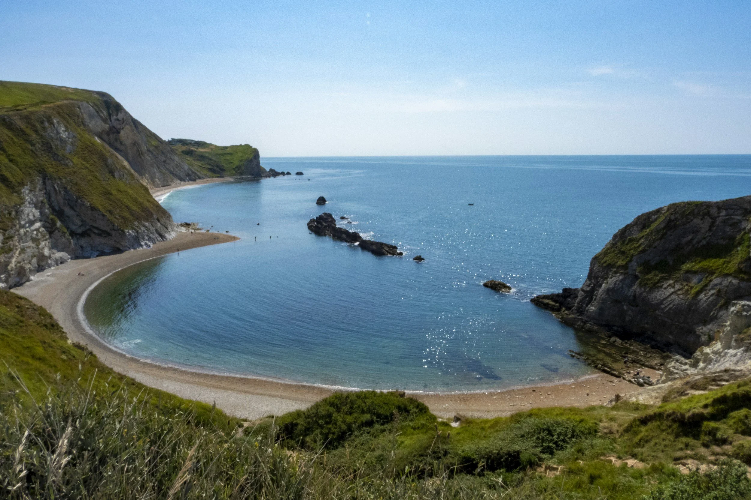 Paysage côtier avec des falaises verdoyantes, une plage en forme de boucle, et l'océan bleu sous un ciel clair.