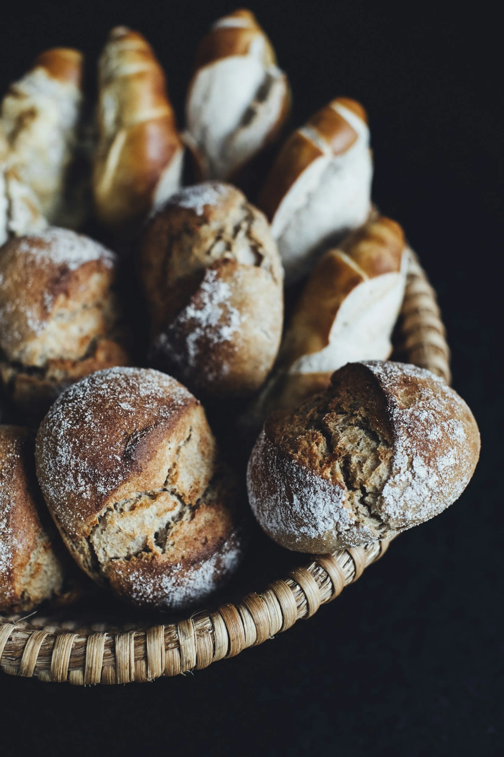 Gros plan de petits pains briochés dans une corbeille en osier, certains saupoudrés de farine, sur un fond noir.