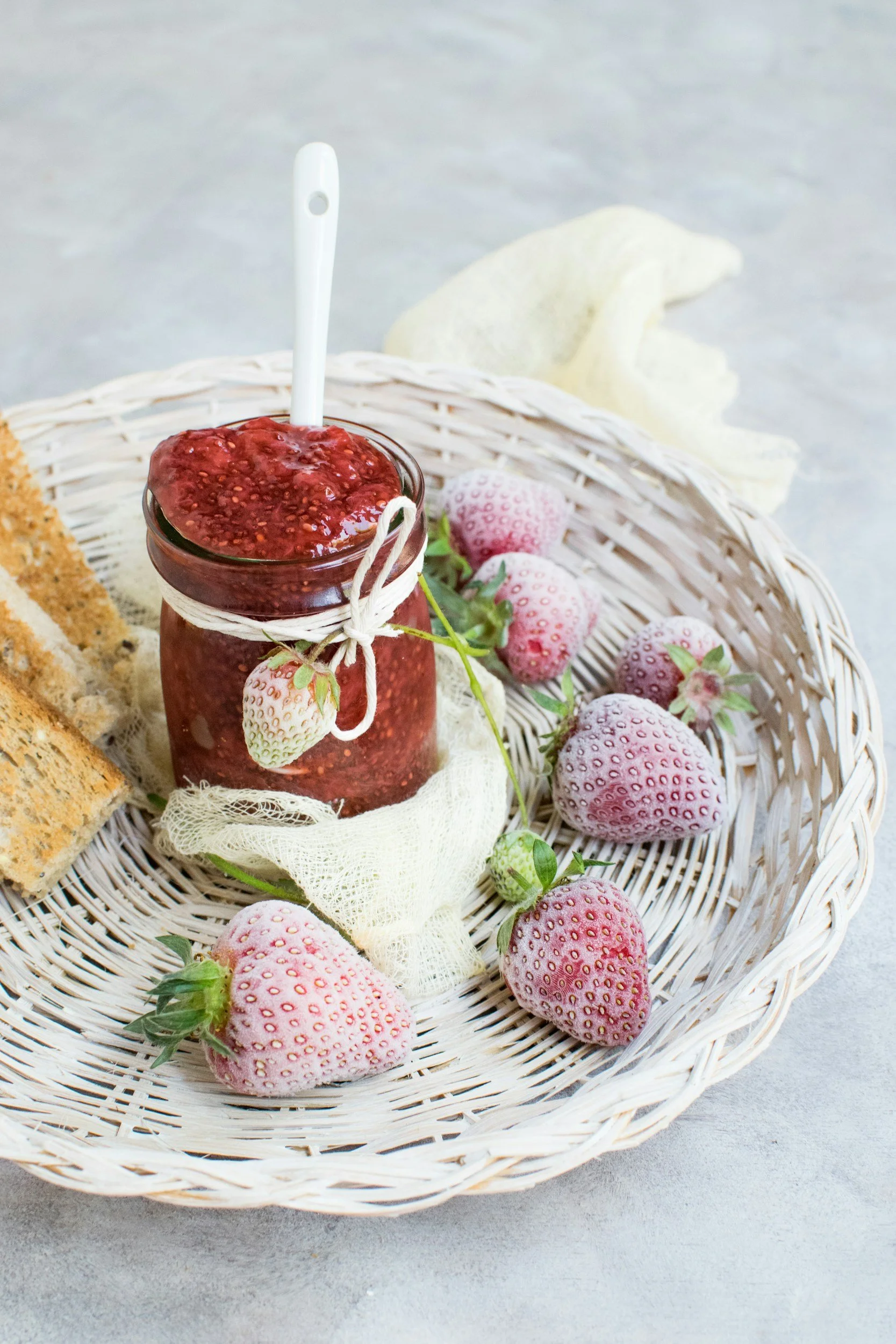 Un panier en osier contenant des fraises, une confiture de fraises en pot avec une cuillère, et des morceaux de gâteau, le tout sur une surface en pierre claire.