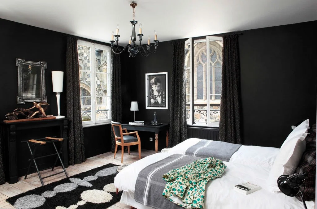 Chambre à coucher avec murs noirs, grand lit avec des draps blancs et une couverture grise, plusieurs fenêtres avec des rideaux foncés, un tapis noir et blanc, un bureau avec une lampe et une chaise en bois, un tableau noir et blanc au mur, un miroir, des décorations, et une robe ou un vêtement à motifs sur le lit.