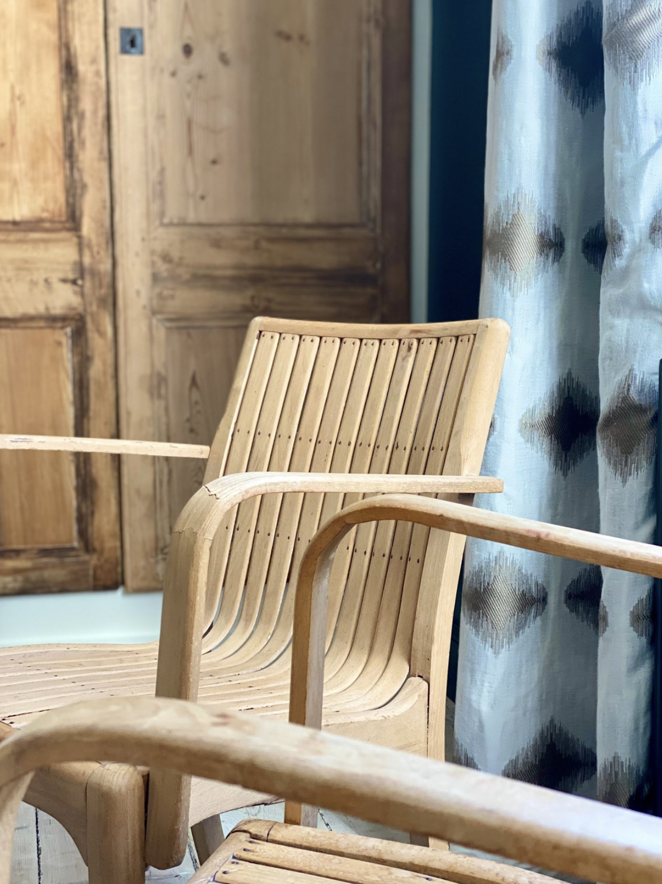 Fauteuil en bois clair avec dossier en lamelles et accoudoirs arrondis, placé à côté de rideaux en tissu avec motif géométrique bleu, blanc et beige, dans une pièce avec mobilier en bois