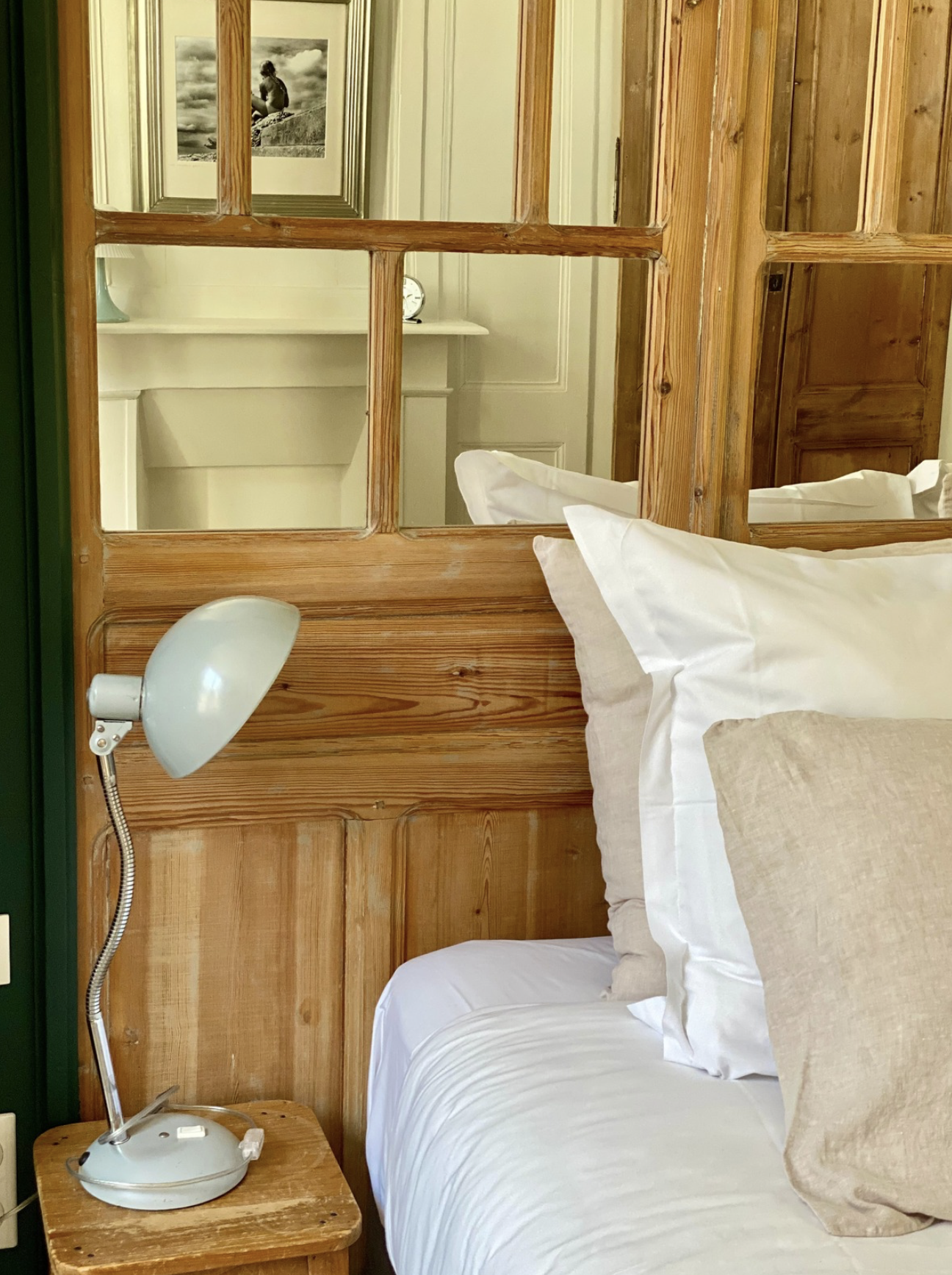 Un lit fait avec des oreillers beige et blanc, avec une tête de lit en bois et une lampe de chevet blanche, vue à travers une cloison en bois avec des petits carreaux de verre.