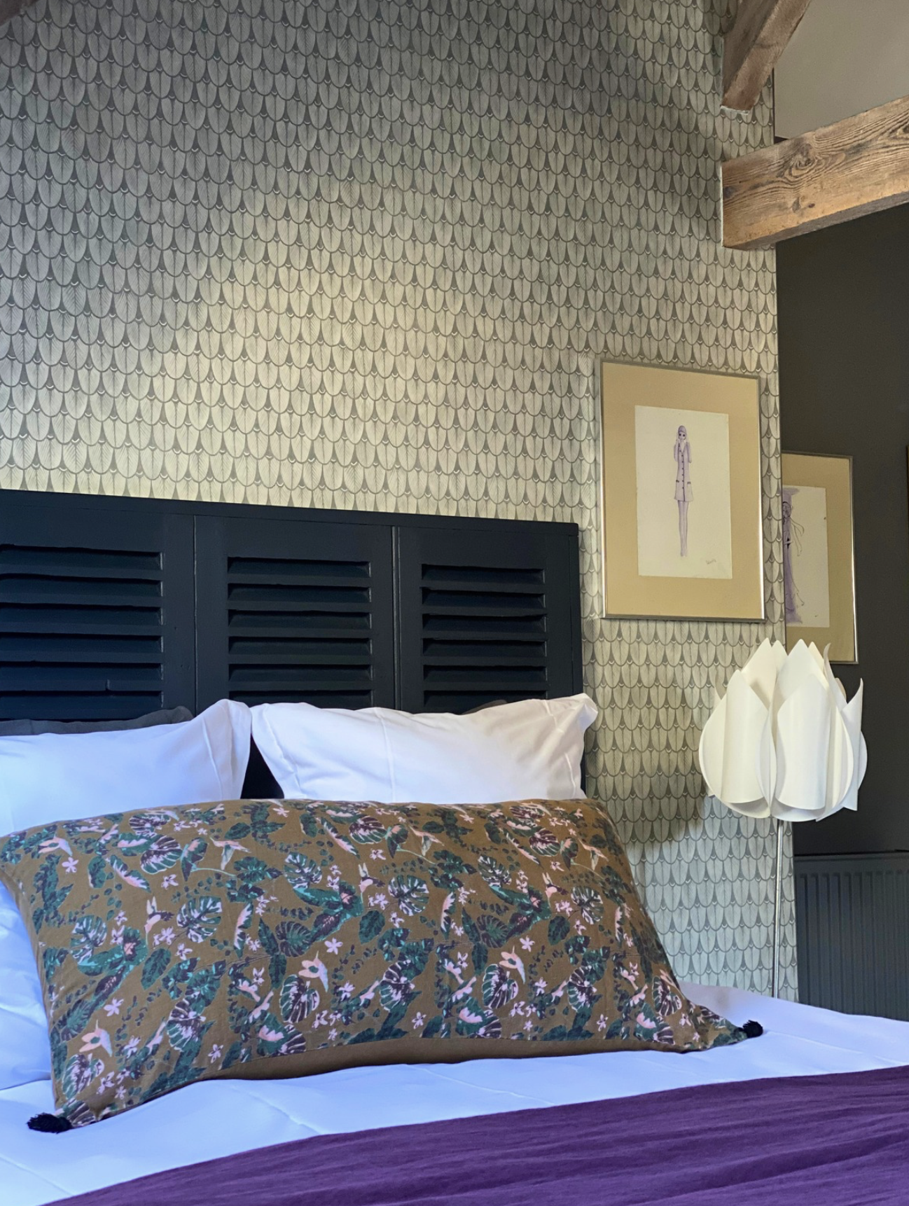Un lit avec des draps blancs, un couvre-lit violet et une taie d'oreiller à motif floral. Au-dessus du lit, un mur décoratif avec un motif d'écailles en beige et gris. Deux tableaux encadrés avec des illustrations de femmes, et un luminaire en forme de fleurs blanches en papier sur le côté.
