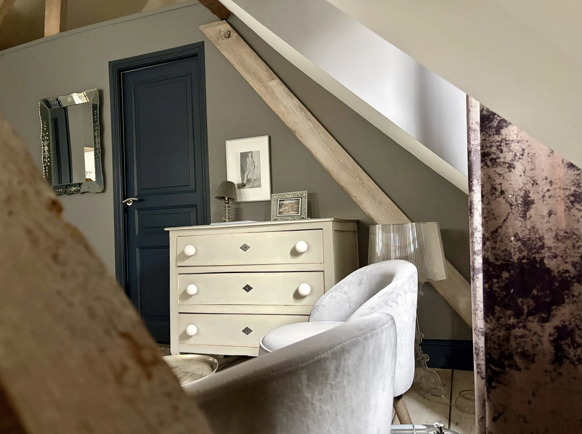 Une pièce d'intérieur avec un mur gris foncé, un miroir avec un cadre orné, une porte en bois peint en bleu marine, un tableau noir et blanc, une lampe de chevet, une commode blanche avec des poignées en losange, un tapis rose et mauve, une table en bois foncé, et un fauteuil en tissu gris clair.
