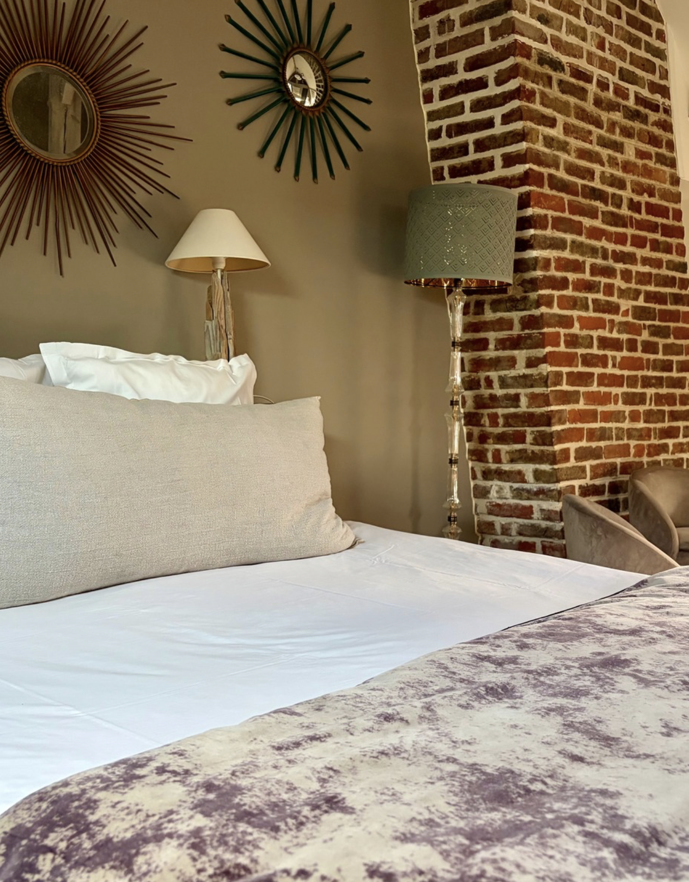 Un lit fait avec des oreillers et une couverture dans une chambre avec un mur en briques et des miroirs décoratifs.