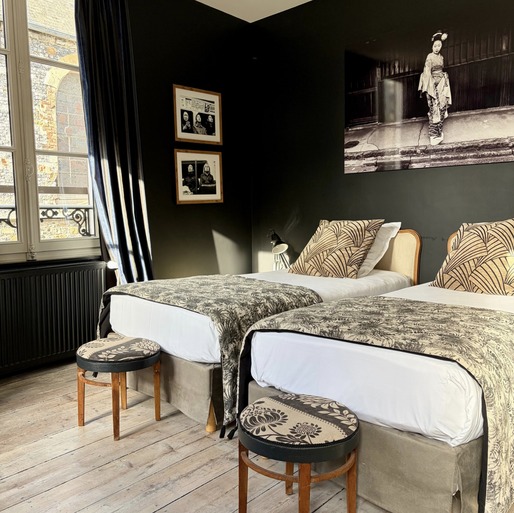 Deux lits simples avec des draps blancs et une couverture à motifs sombre, accompagnés de deux petits tabourets avec coussins à motifs floraux, dans une chambre avec murs gris foncé et un parquet en bois clair. Sur le mur, une grande photographie en noir et blanc représentant une femme en kimono traditionnel japonais. À côté, deux cadres avec des photos en noir et blanc, dont une avec trois femmes.