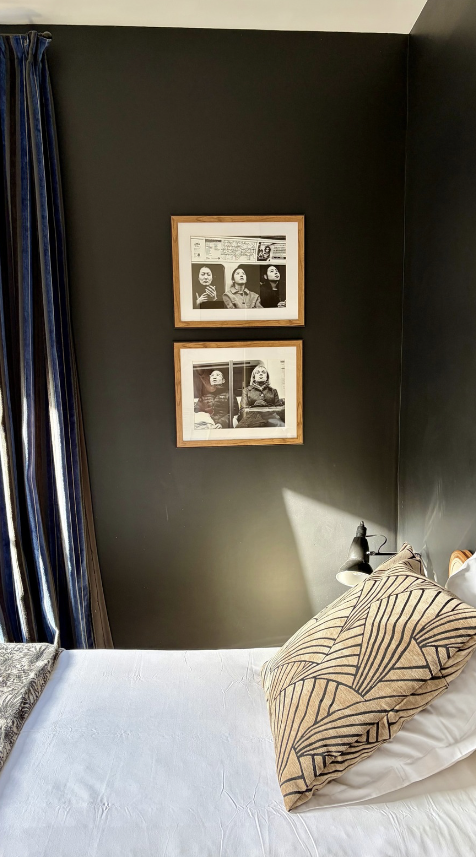 Coin calme d'une chambre avec deux cadres de photos en noir et blanc accrochés au mur gris, un lit avec des draps blancs, une lampe de lecture noire, et un coussin beige avec un motif noir sur le lit.