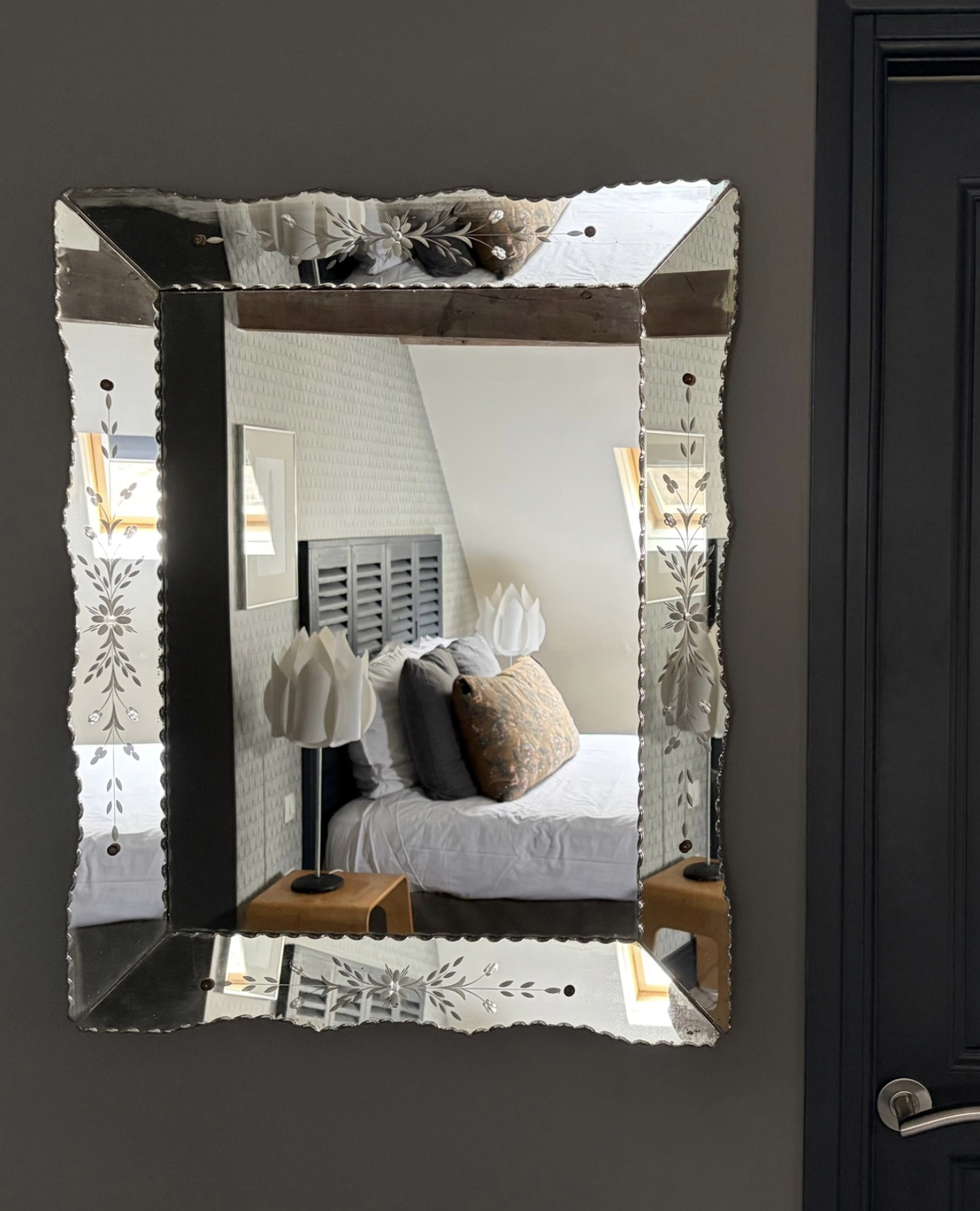 Un miroir orné de détails en métal, reflétant une chambre avec un lit, des lampes de chevet, un tableau et des fenêtres avec des rideaux jaunes.