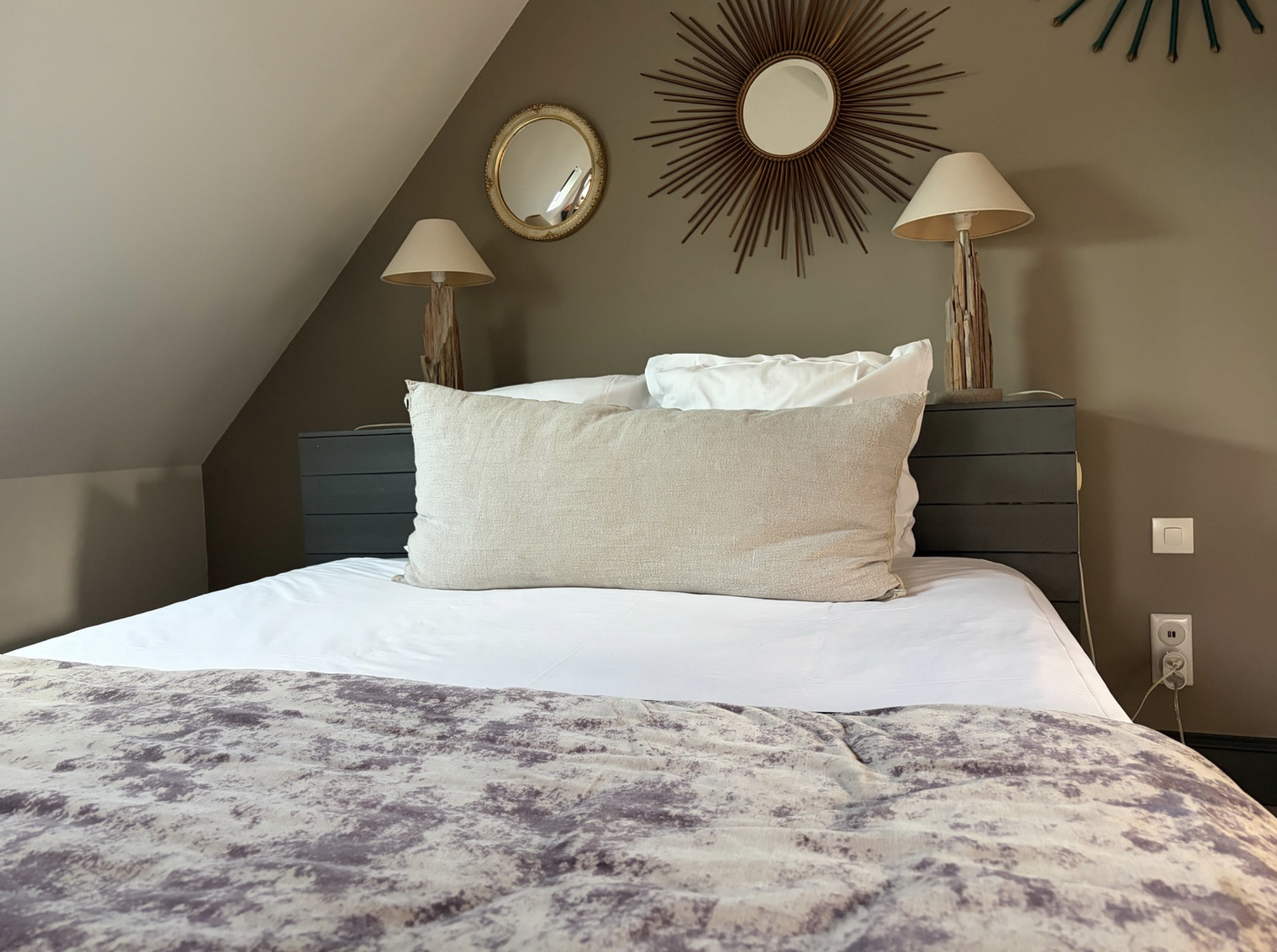 Une chambre avec un lit, deux lampes de chaque côté, deux miroirs sur le mur avec décorations radiantes, et une prise électrique visible à droite.