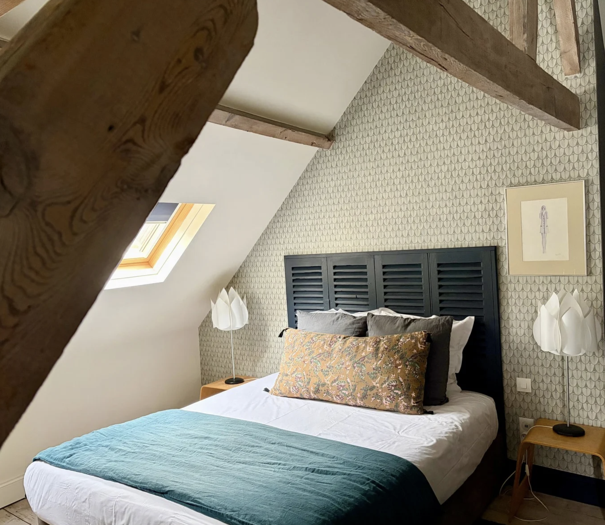 Chambre avec lit fait, oreillers, lampe de chevet, fenêtres de toit, tableau d'art sur le mur, rideaux aux fenêtres.
