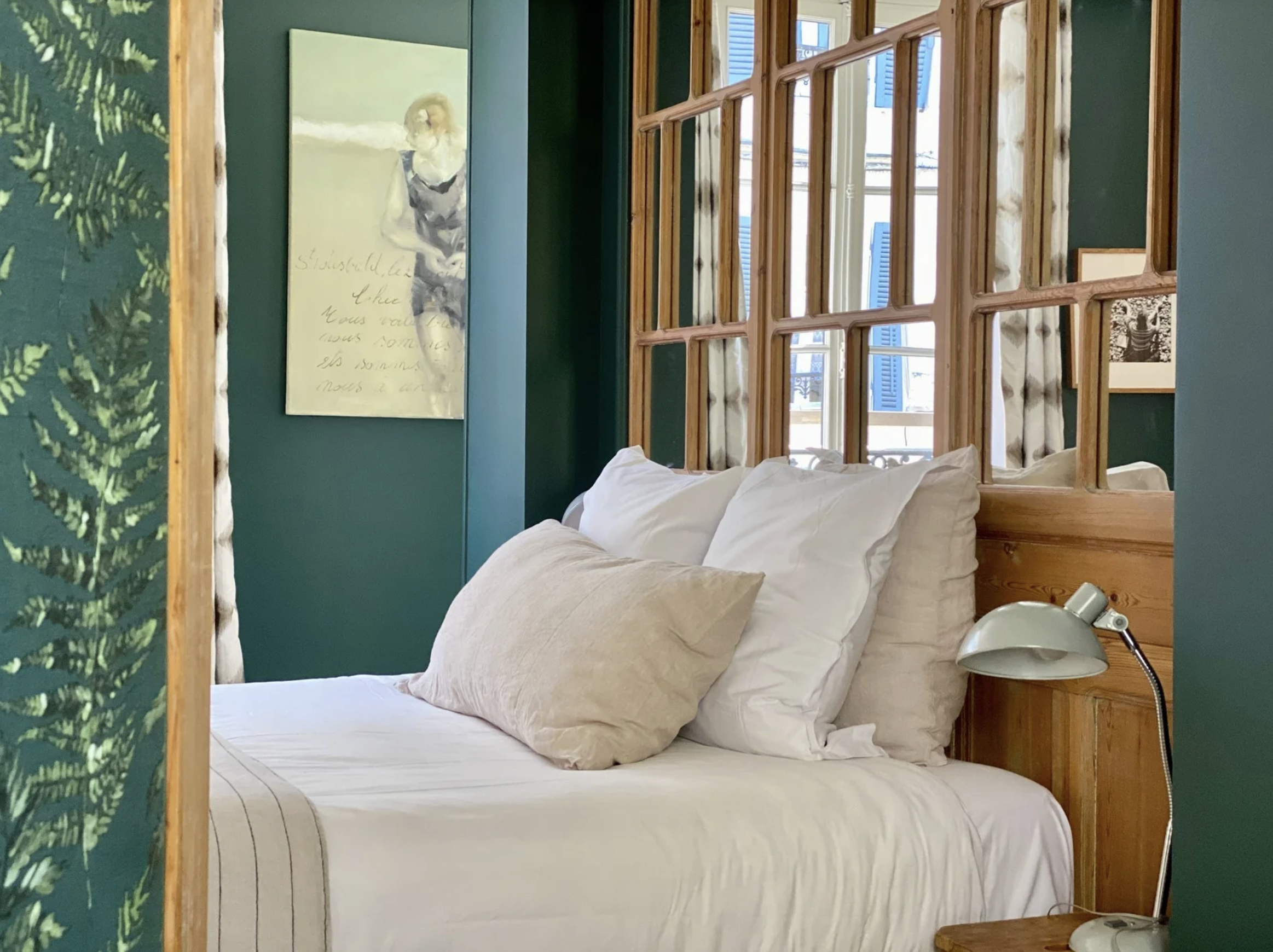 Un lit avec des draps blanc cassé et deux coussins, une lampe de bureau à côté, un mur vert avec une peinture et un miroir en bois, une fenêtre avec des volets bleus.
