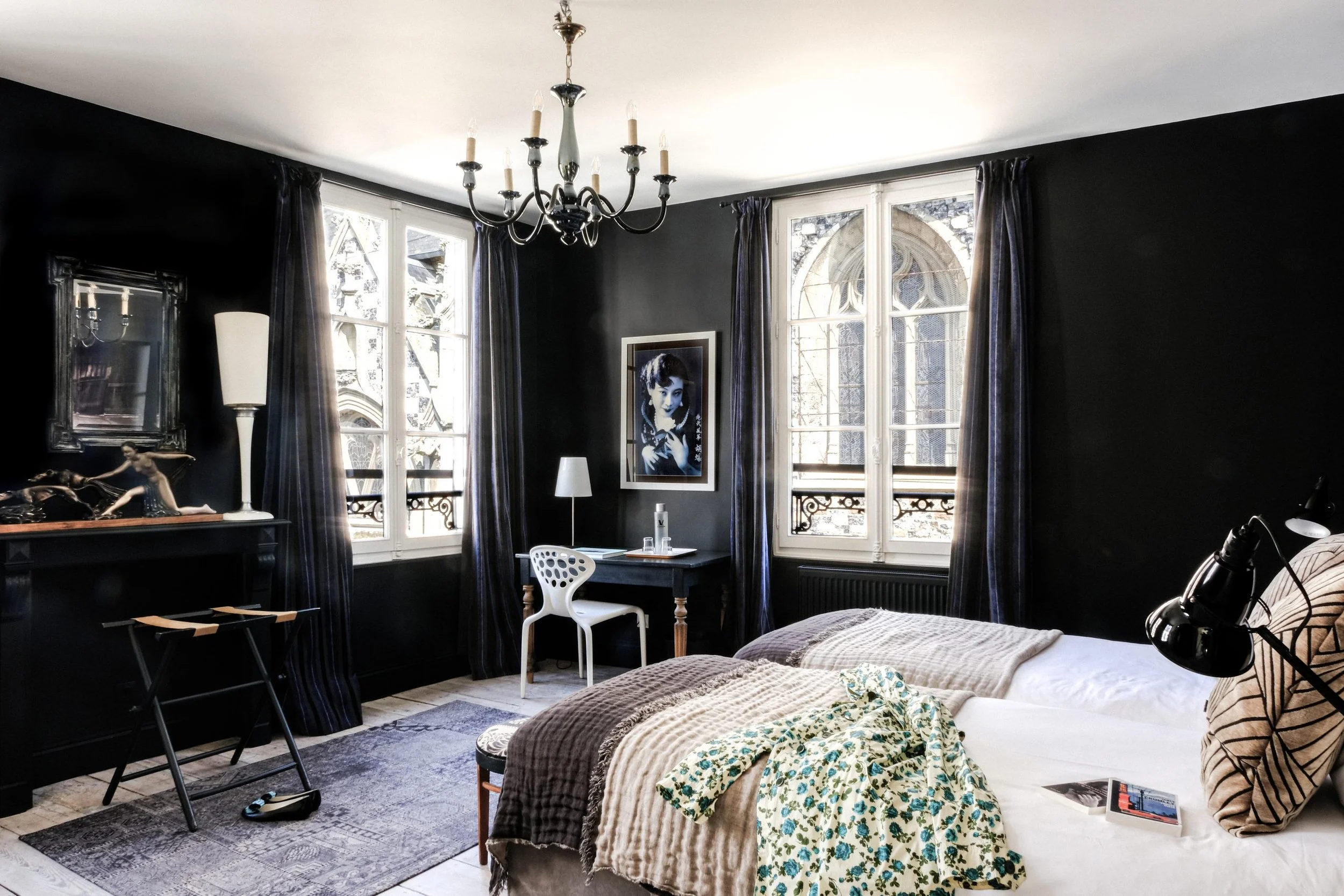 Chambre avec murs noirs, grande fenêtre avec rideaux violets, portrait en noir et blanc d'une femme sur le mur, chandelier noir suspendu, lampe à pied blanche, lit avec une couverture beige, objets décoratifs, miroir et tableau