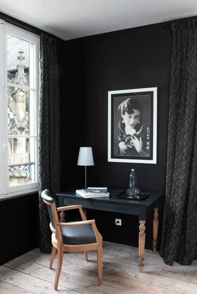 Intérieur d'un bureau avec un mur noir, une fenêtre avec rideaux noirs, un tableau noir et blanc d'une femme âgée, une table avec une bouteille d'eau, des verres, une lampe blanche, un livre, un clavier, une chaise en bois avec assise en cuir noir.