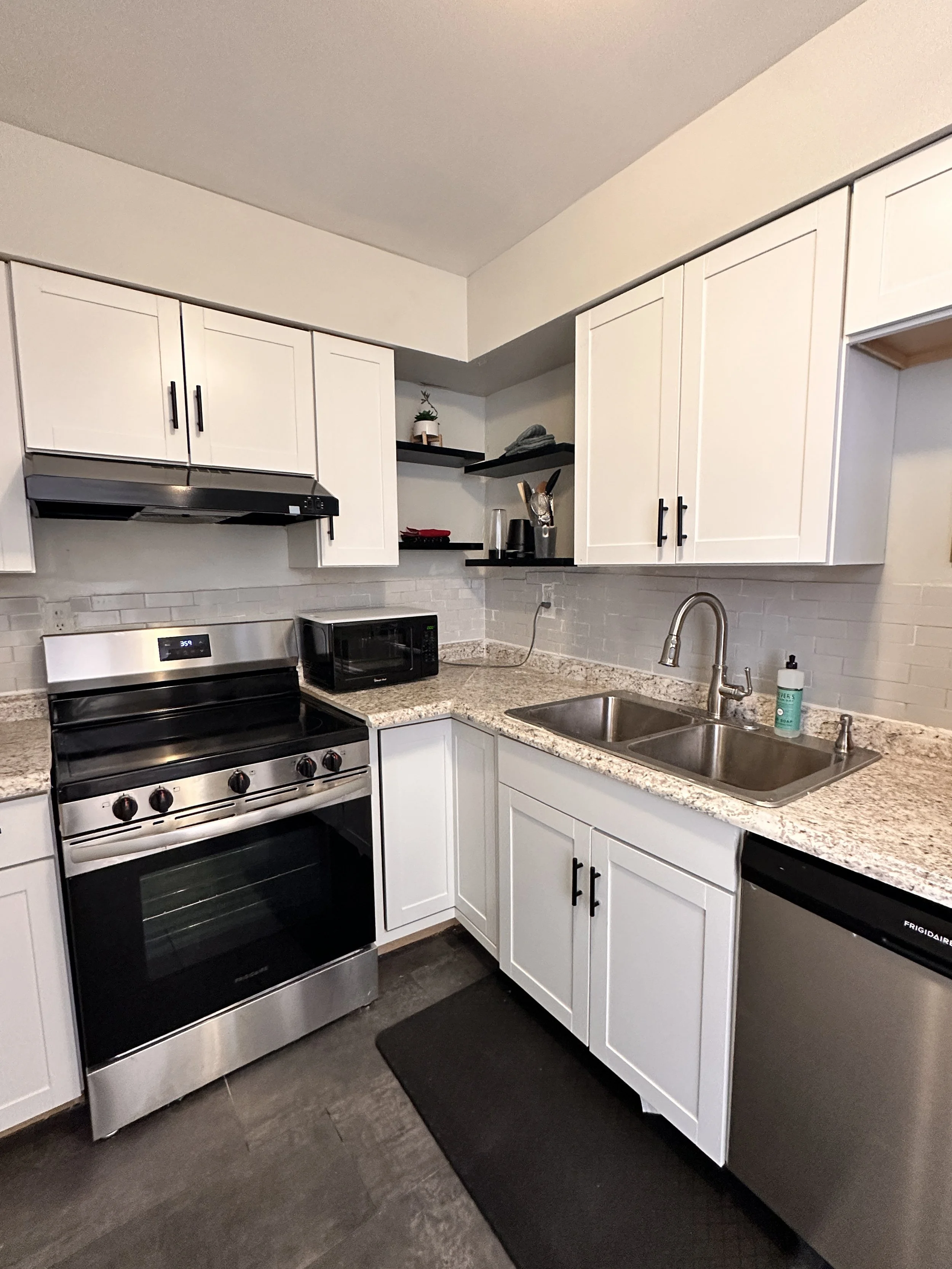 Kitchen with white cabinets, stainless steel stove, microwave, and dishwasher, granite countertops, white subway tile backsplash, double sink, and black open shelves with small decor items.