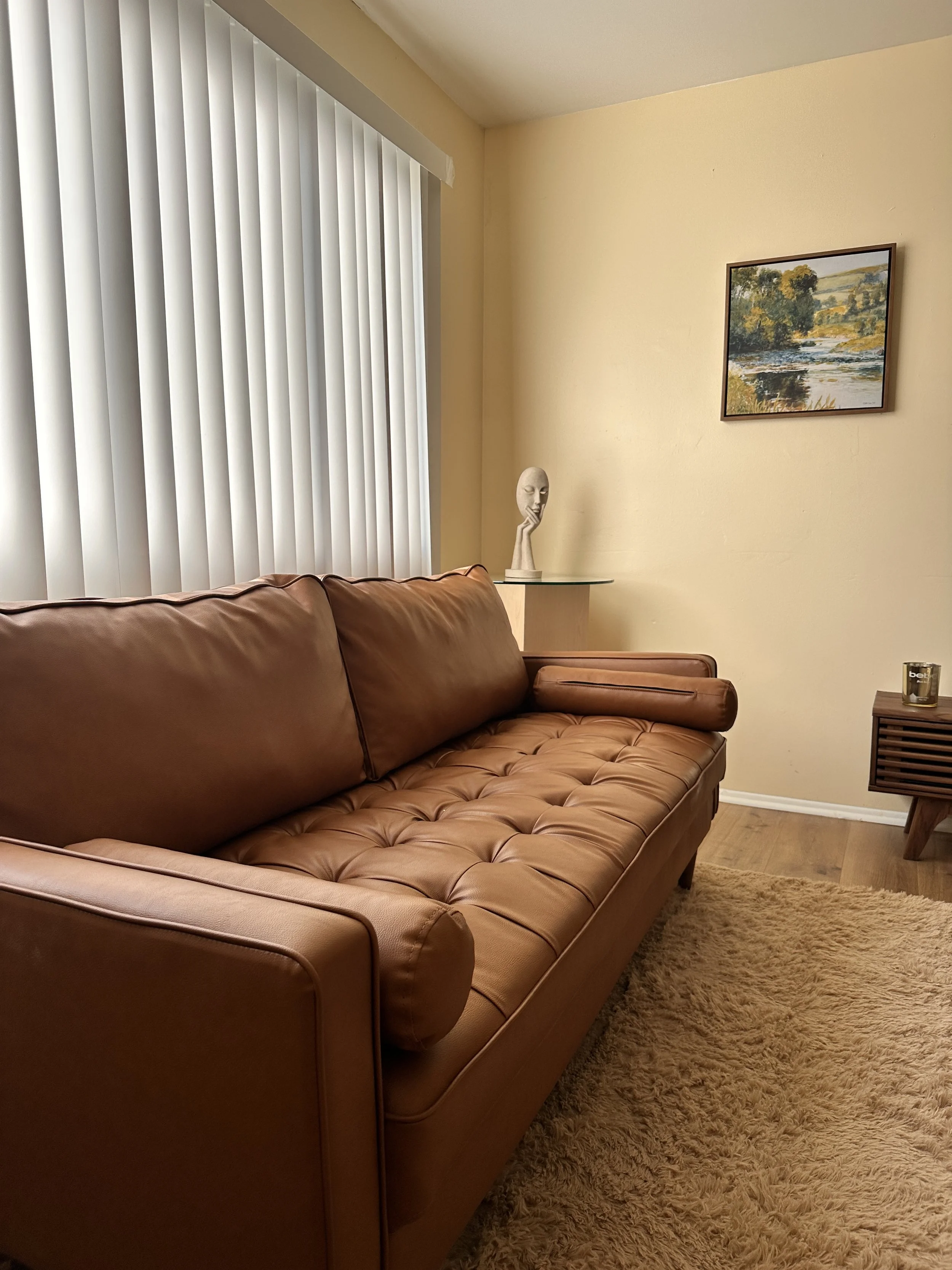 A living room with a brown leather tufted sofa, a window with white vertical blinds, a round side table with a white sculpture of a face resting on a hand, a framed landscape painting on the wall, and a beige shaggy rug.