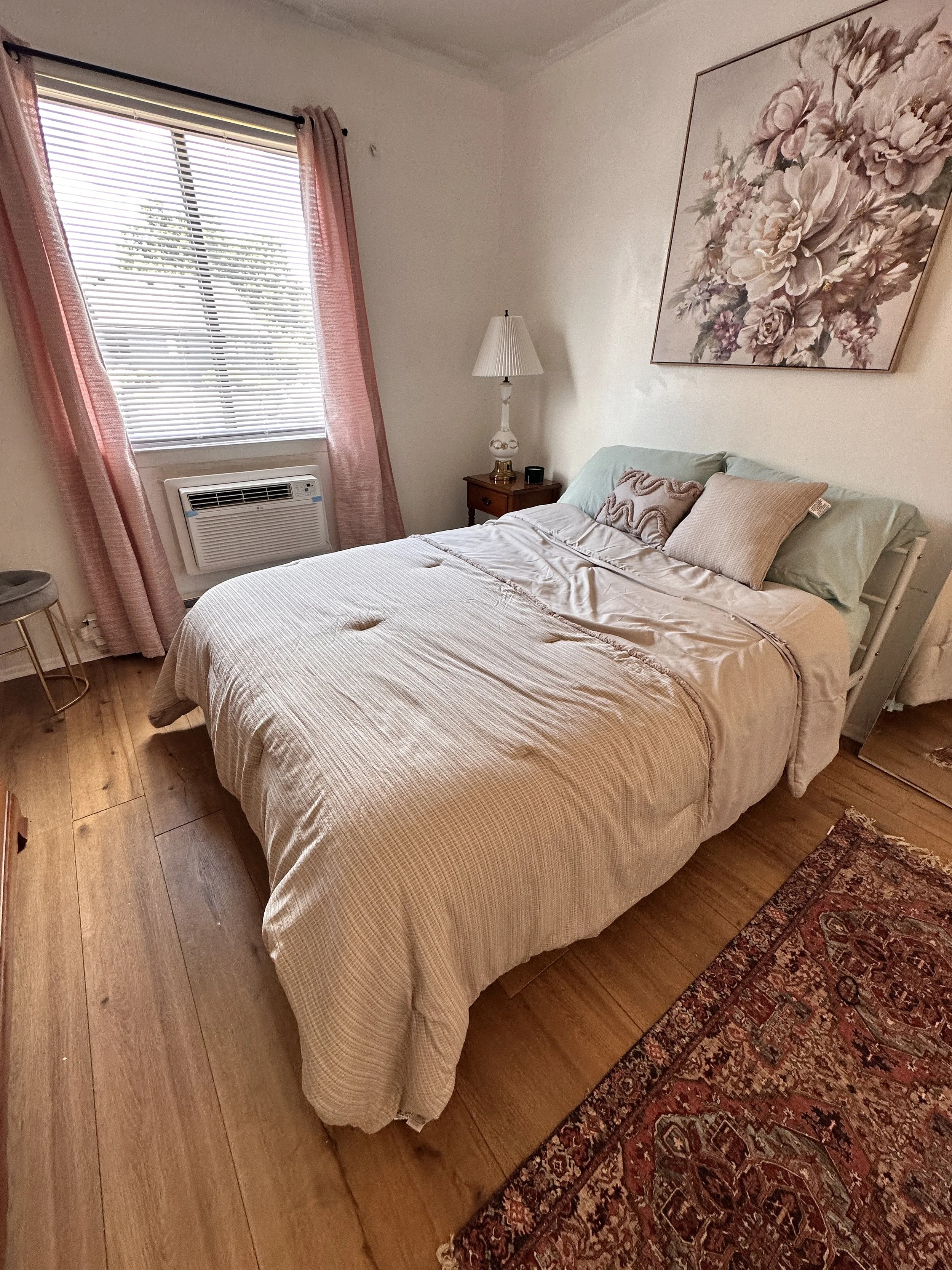 A bedroom with a bed, a nightstand with a lamp, a window with pink curtains, a floral painting, a small stool, and a patterned rug.