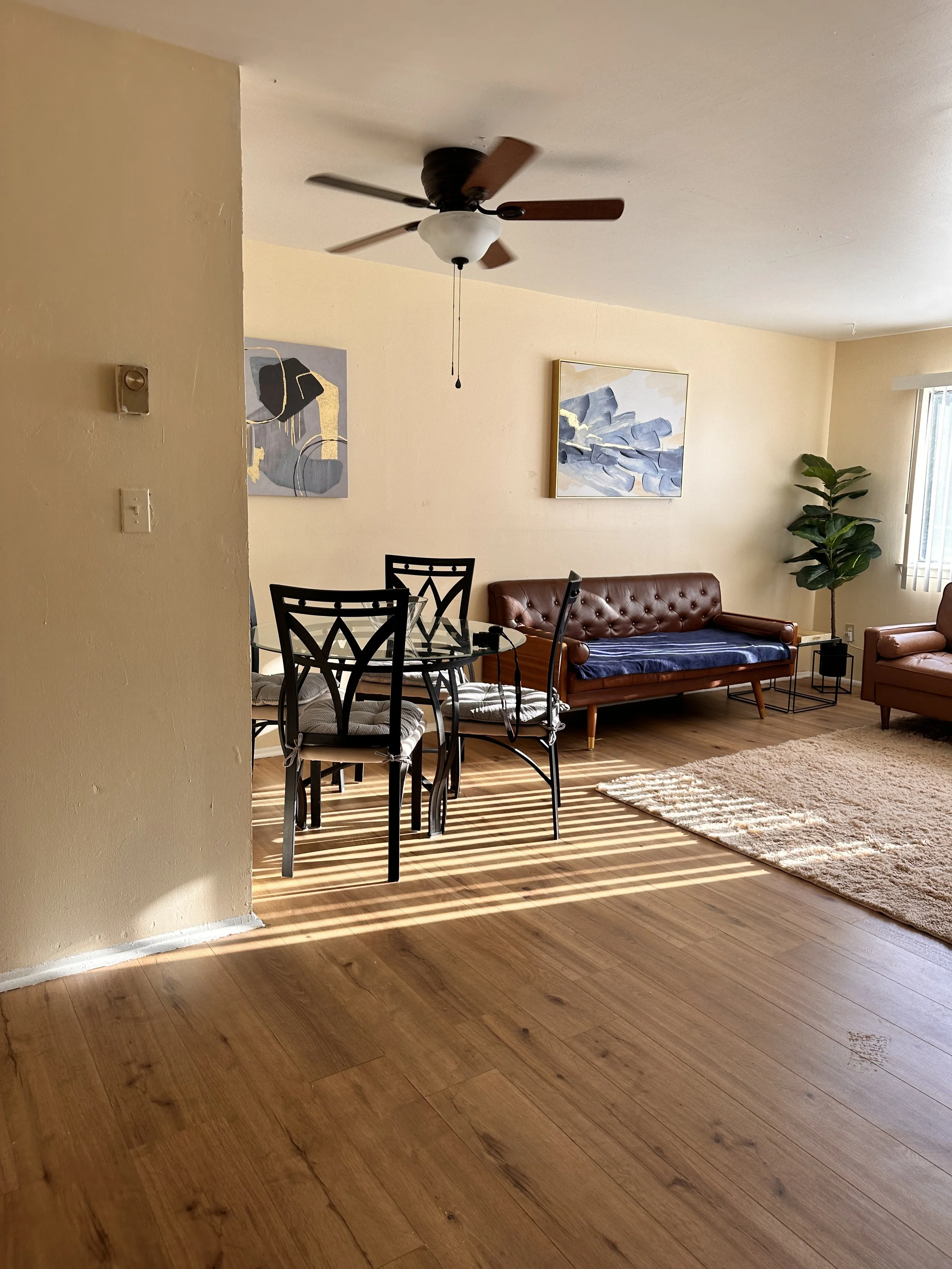 Living room with a ceiling fan, wooden flooring, a brown leather sofa, a small armchair, a glass dining table with four black chairs, artwork on the walls, a potted plant, and a window with vertical blinds.