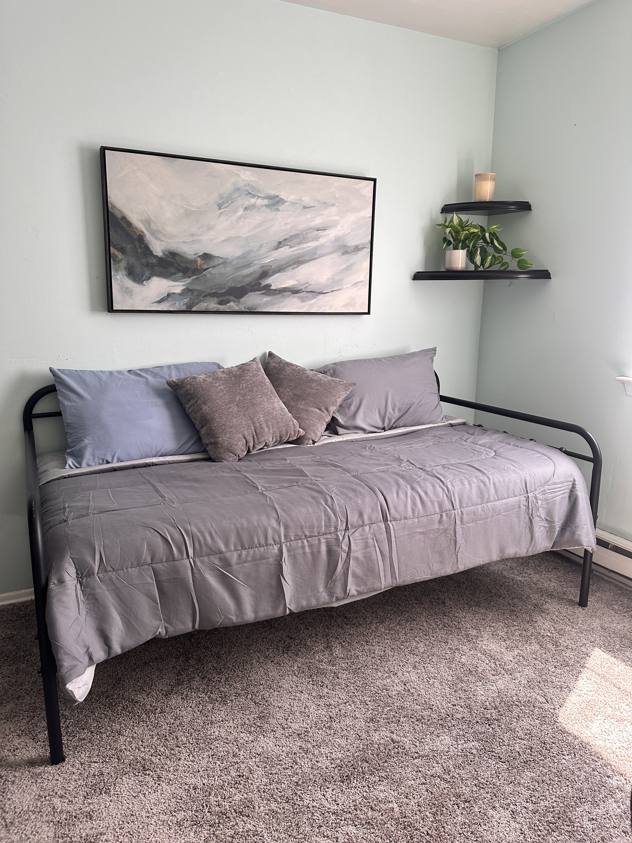 A bedroom with light blue walls, a black metal bed frame, and gray bedding with matching pillows. There are two gray pillows and two darker gray pillows on the bed. Above the bed is an abstract painting with shades of gray, white, and black. To the r