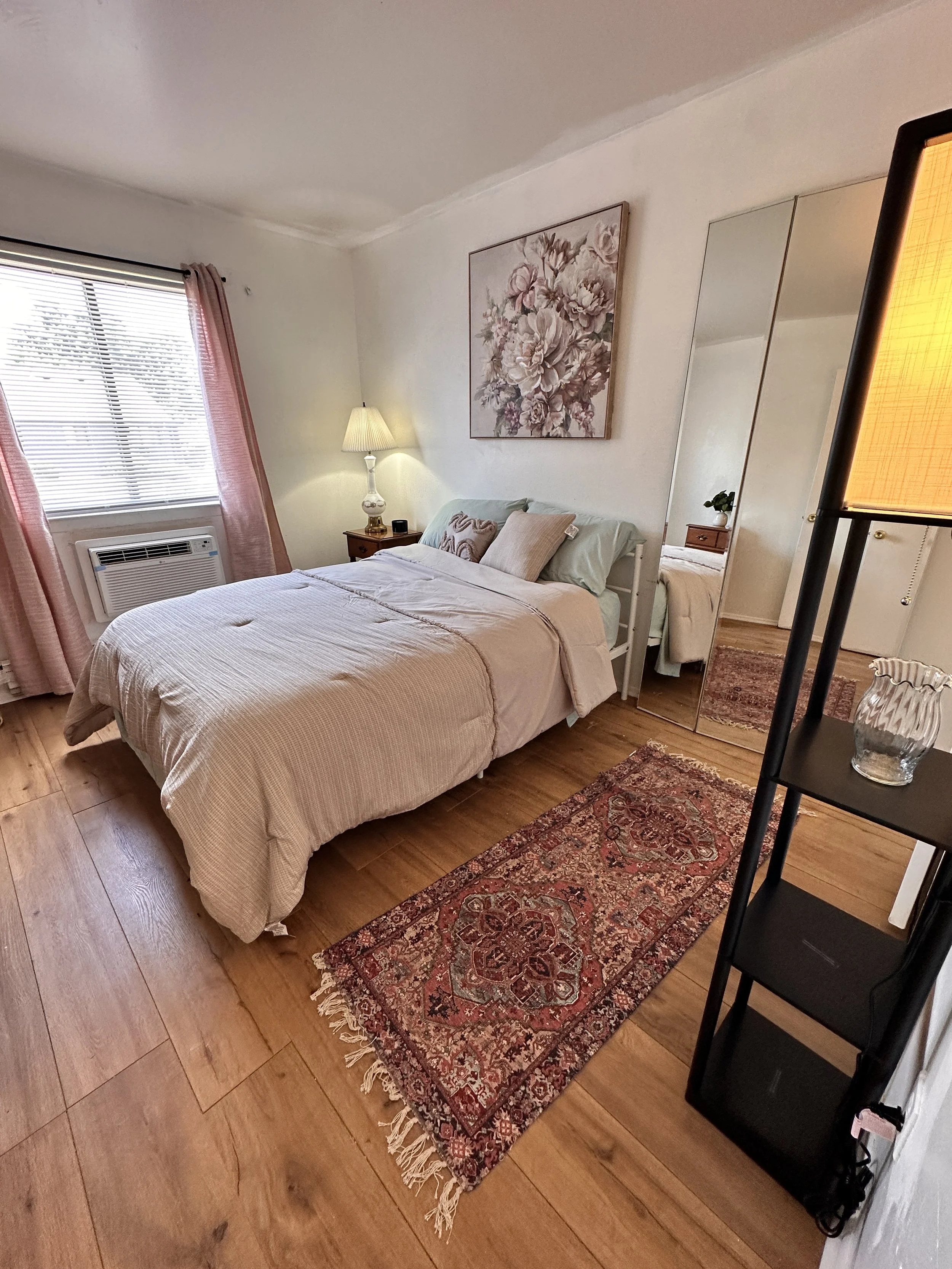 A cozy bedroom with a bed, a bedside table with a lamp, a mirror, a window with pink curtains, a colorful rug, and a decorative painting of flowers on the wall.
