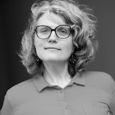 Black-and-white portrait of a woman with curly hair, glasses, and earrings wearing a collared shirt.
