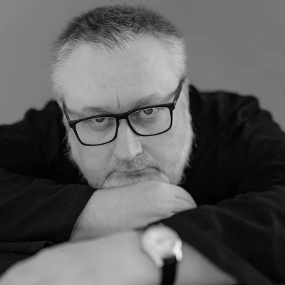Black and white close-up portrait of a middle-aged man with short hair, beard, glasses, resting his head on his folded arms, looking at the camera.