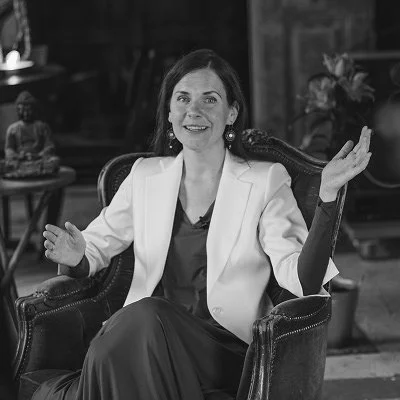 A woman sitting in a vintage-style armchair, smiling, wearing a white blazer and dark dress, with plants and decorative items in the background.