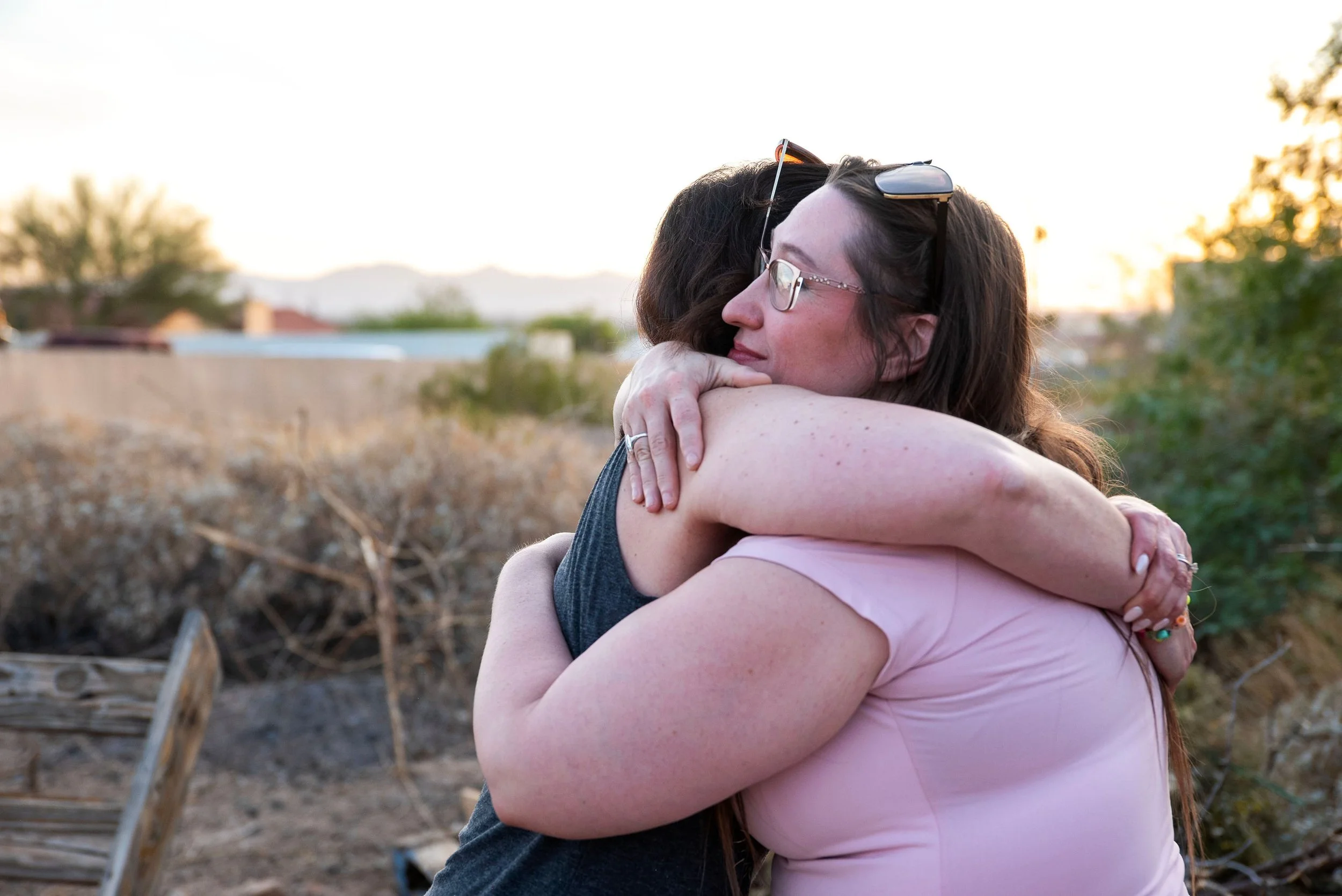Two women hugging outdoors during sunset, one with dark hair, glasses, and sunglasses on her head, the other with light skin, long hair, and a pink top, in a natural setting with trees and a fence in the background.