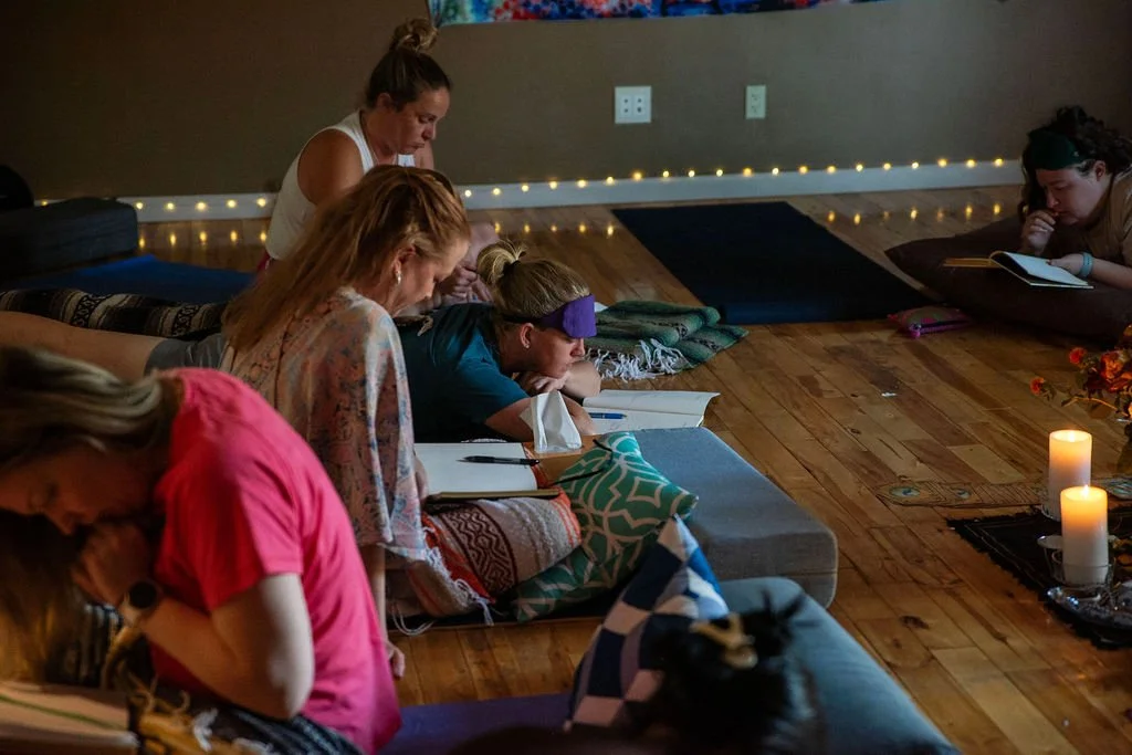 A group of women and girls participating in a meditation or prayer session indoors on a wooden floor with candles and string lights in the background.