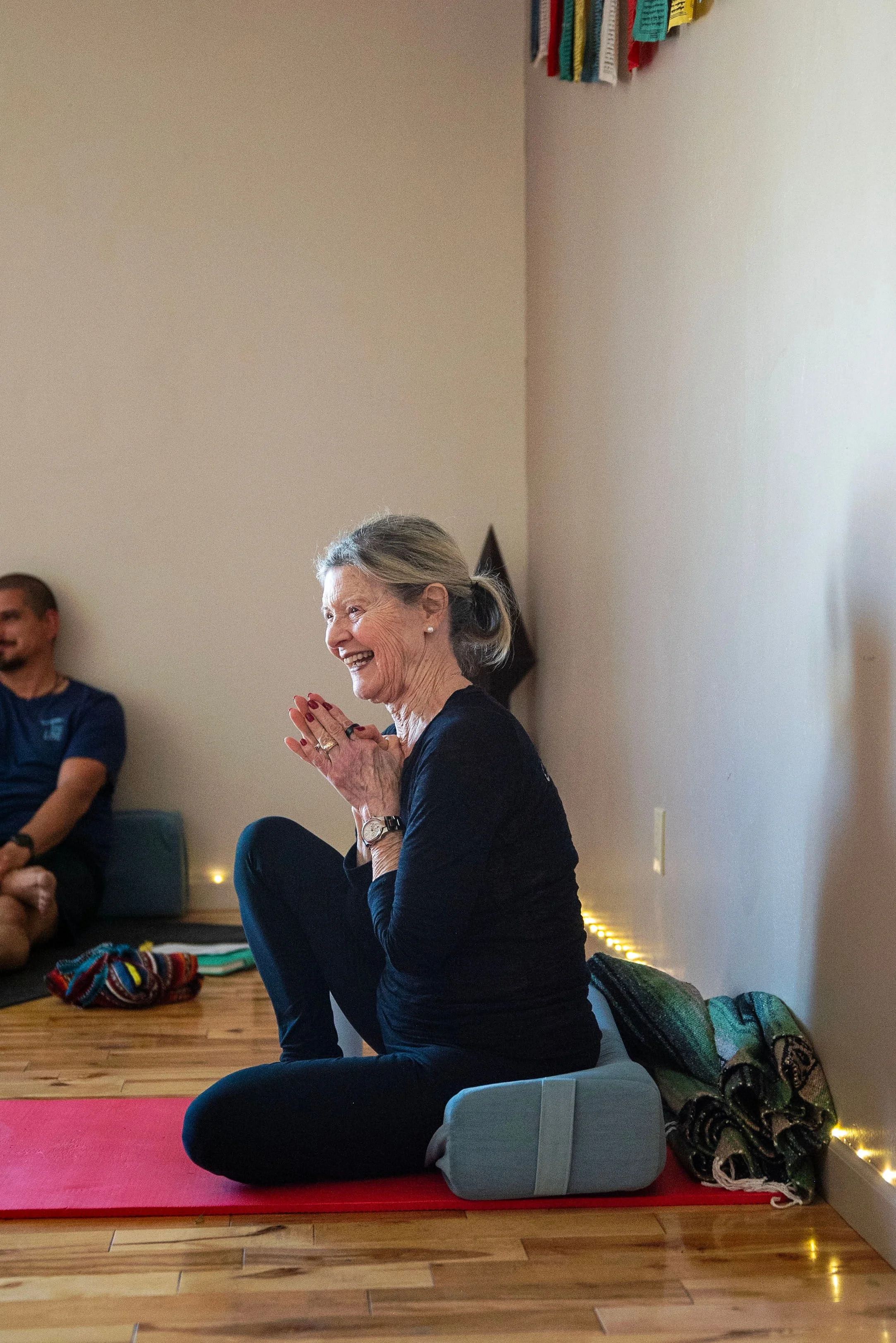 An older woman is sitting on a yoga bolster, smiling and clapping her hands together in a seated yoga or meditation practice. She is wearing a black long sleeve shirt and black pants, with a watch on her left wrist. There is a colorful blanket next to her on the floor. Other people are seated nearby; one in a blue shirt is partially visible in the background. The room has wooden floors, neutral-colored walls, and string lights along the baseboard.