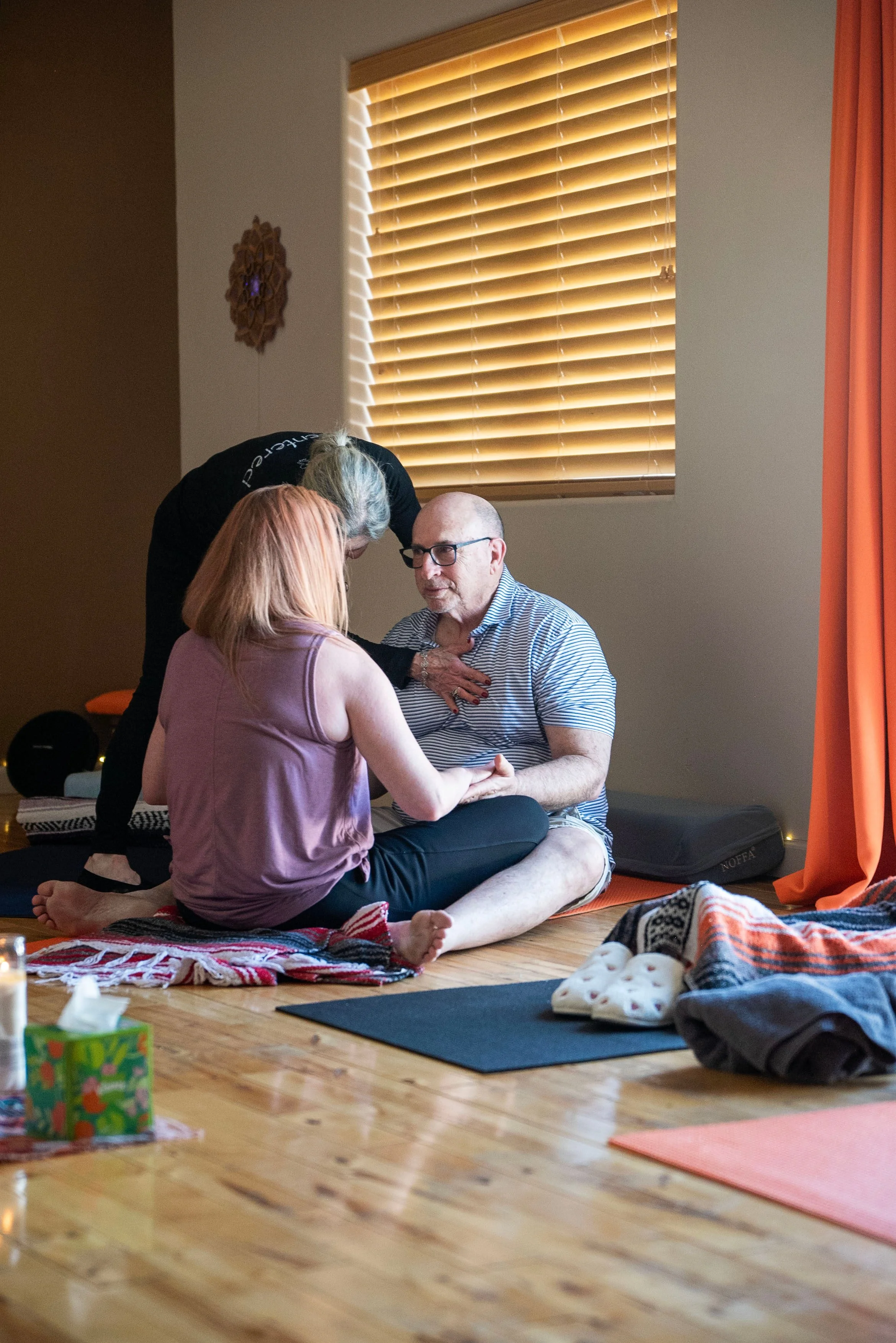 A group of three people practicing yoga or meditation indoors, sitting on a colorful blanket on a wooden floor, with a window with closed blinds and orange curtains in the background.