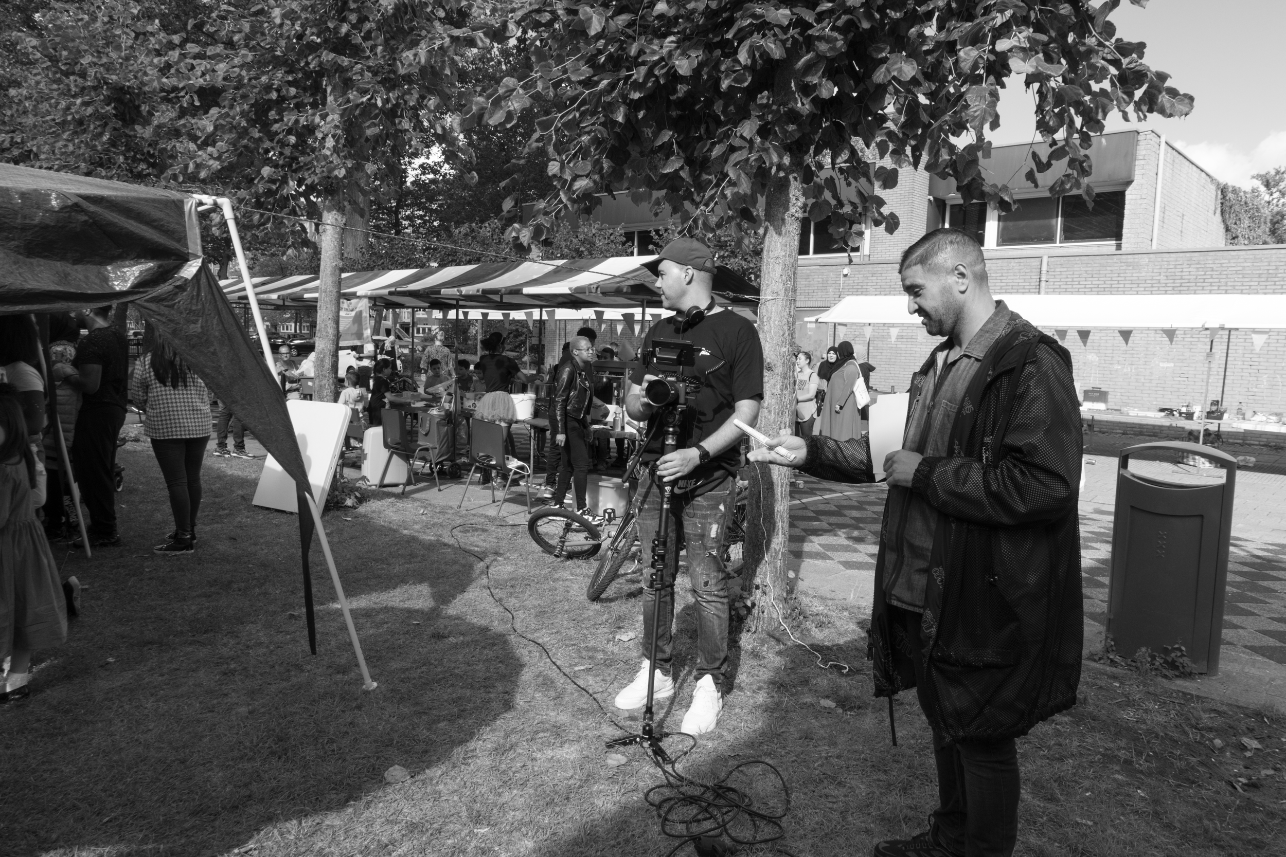 Two men are standing outdoors near trees and a street market. One man is holding a camera on a tripod, and the other man is looking at his phone. There are people and tents in the background, and a building behind them.