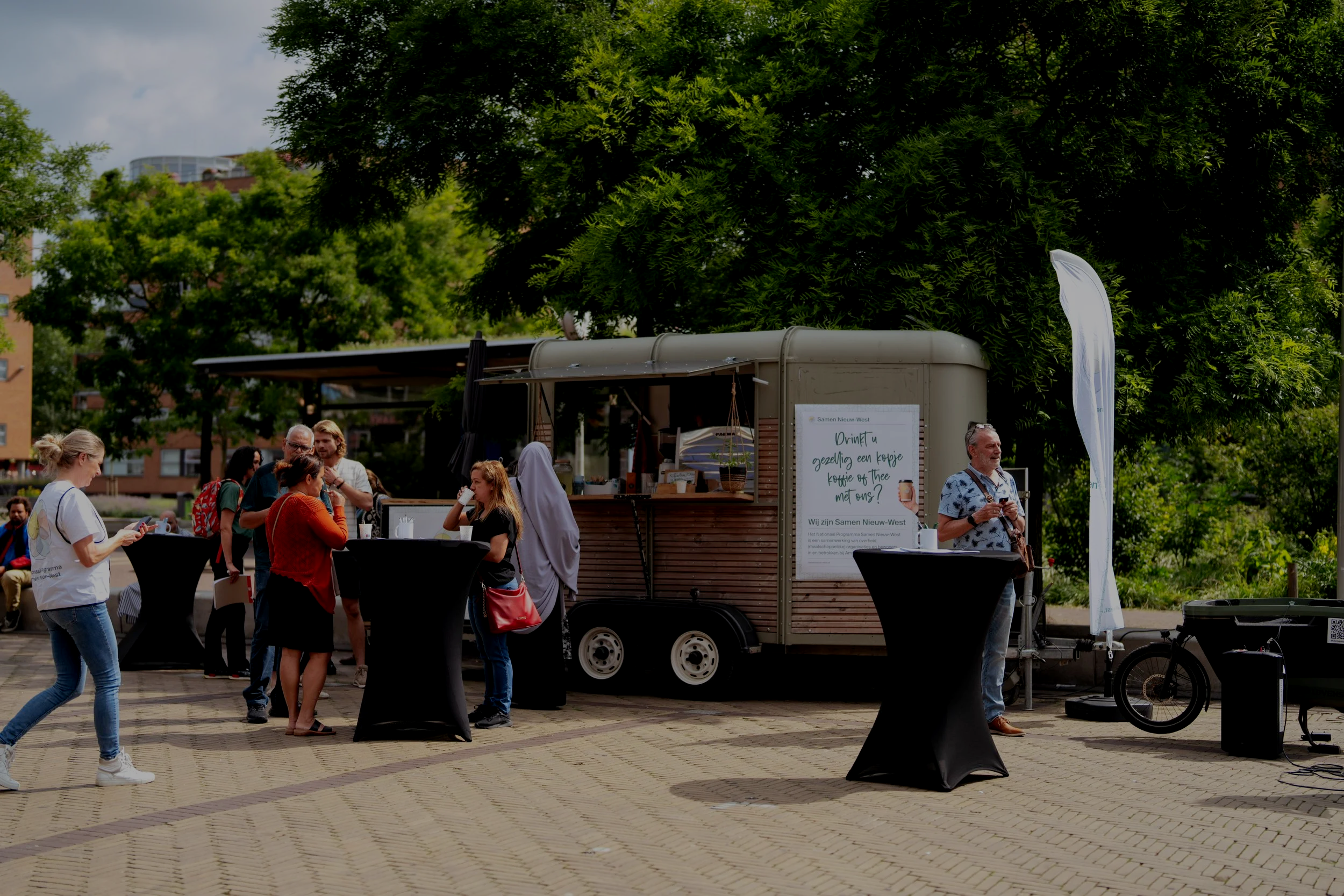 People gathering outdoors around a food truck with tall black cocktail tables, with trees and city buildings in the background.