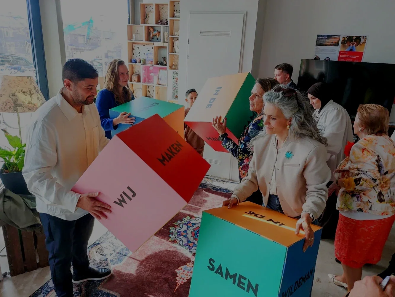 Group of people with large colorful blocks in a room with a bookshelf, a television, and large windows.