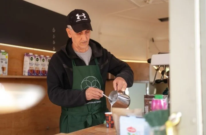 An elderly man wearing a black cap and a green apron pouring a liquid into a cup at a coffee stand or cafe.