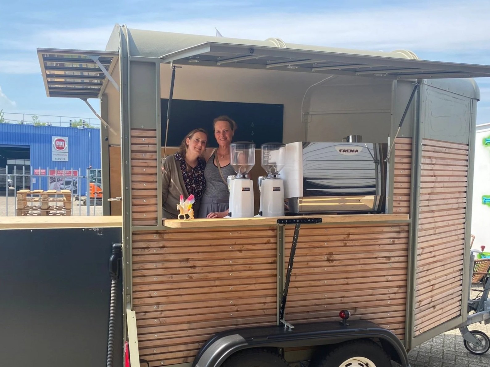 Two women inside a mobile coffee cart, smiling at the camera, with coffee machines and a small decorative unicorn on the counter. The cart is made of wood and used for selling beverages.