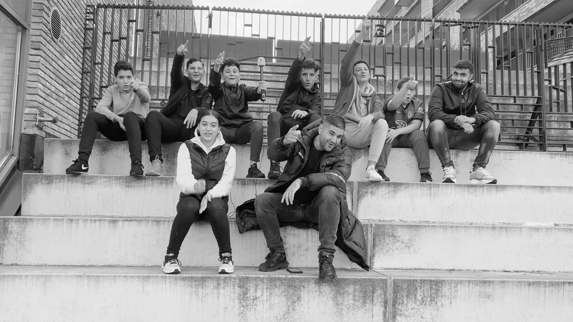 Group of nine children and two adults sitting on outdoor concrete steps with a metal fence behind them. The children are in casual clothes, some raising their hands and pointing, one holding an umbrella. The adults are smiling, one making a gesture, and the other resting with legs crossed. The scene is in black and white.