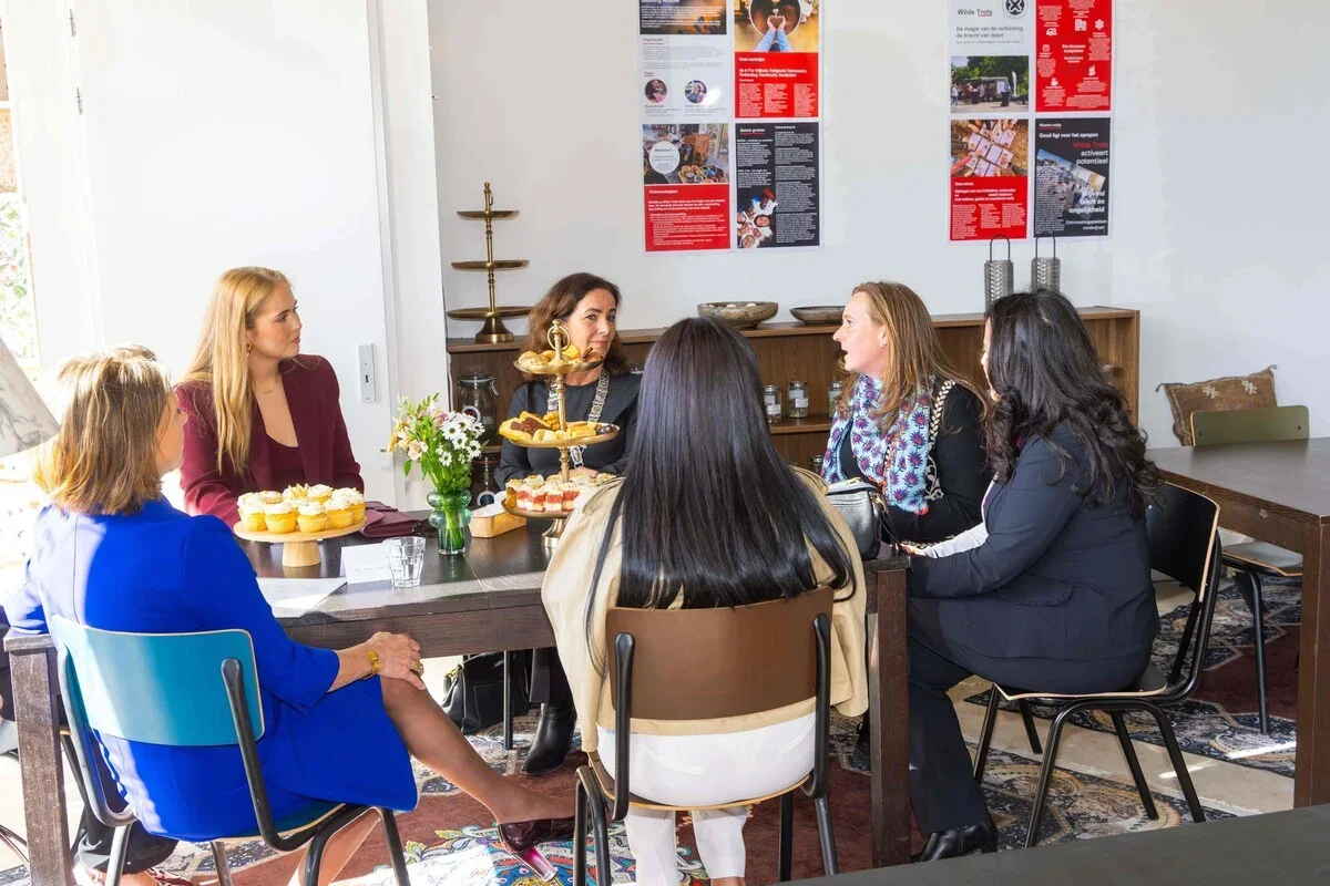 Six women sitting around a table in a bright room, having a discussion. The table is decorated with a vase of flowers and a tray of cupcakes. The background shows posters on the wall and a wooden sideboard.