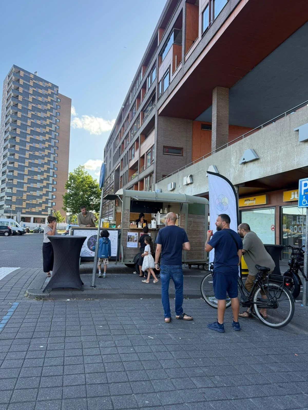 People gathered around a street food kiosk in an urban area, with high-rise buildings in the background on a sunny day.